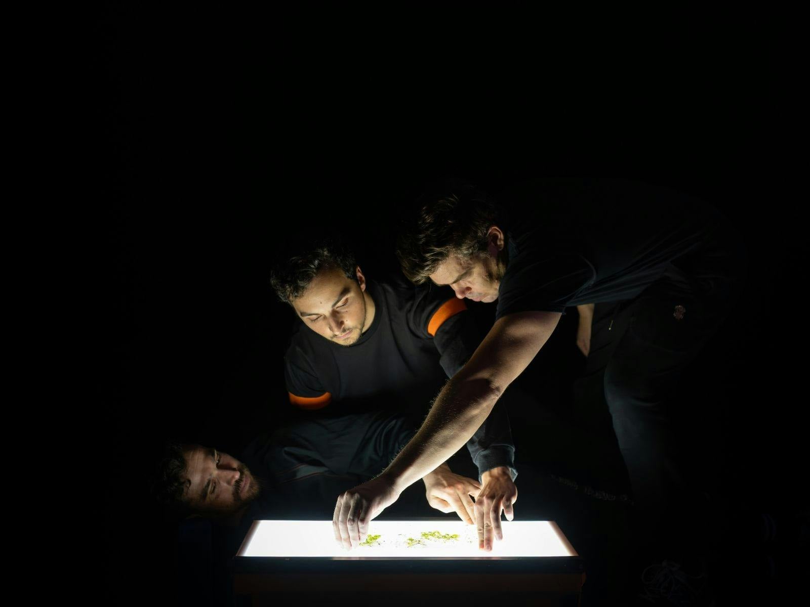 Three men wearing black and their arms interlinked looking over a lightbox