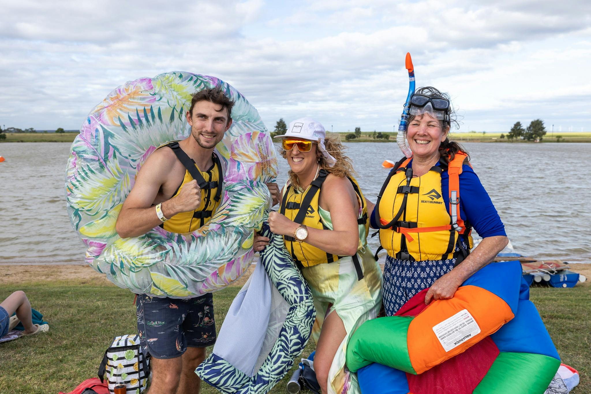 A group of three people with inflatable water rings . They are wearing lifejackets and lakeside