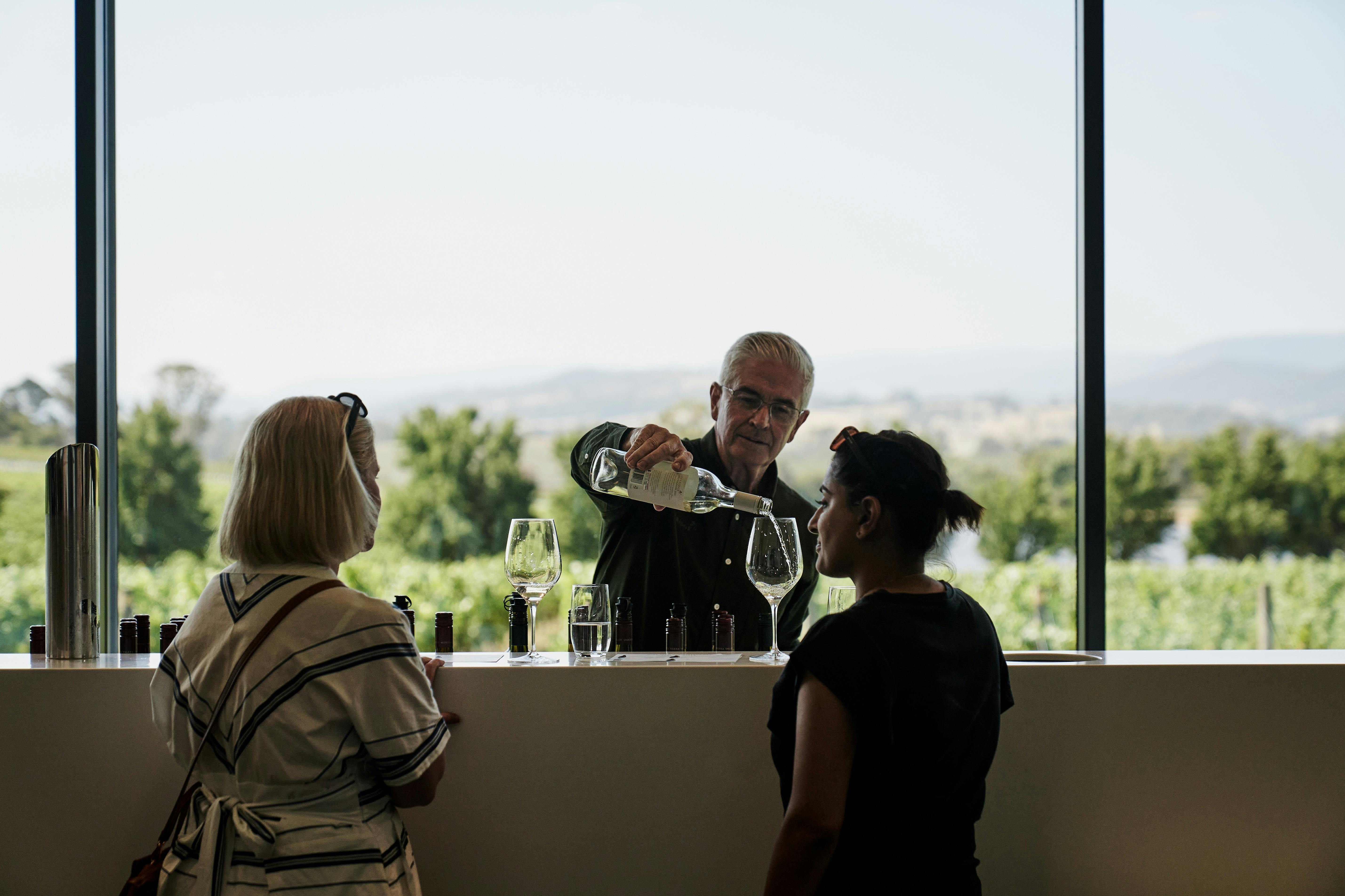 2 women are seated at Oakridge's bar with a view to the vineyard beyond. A