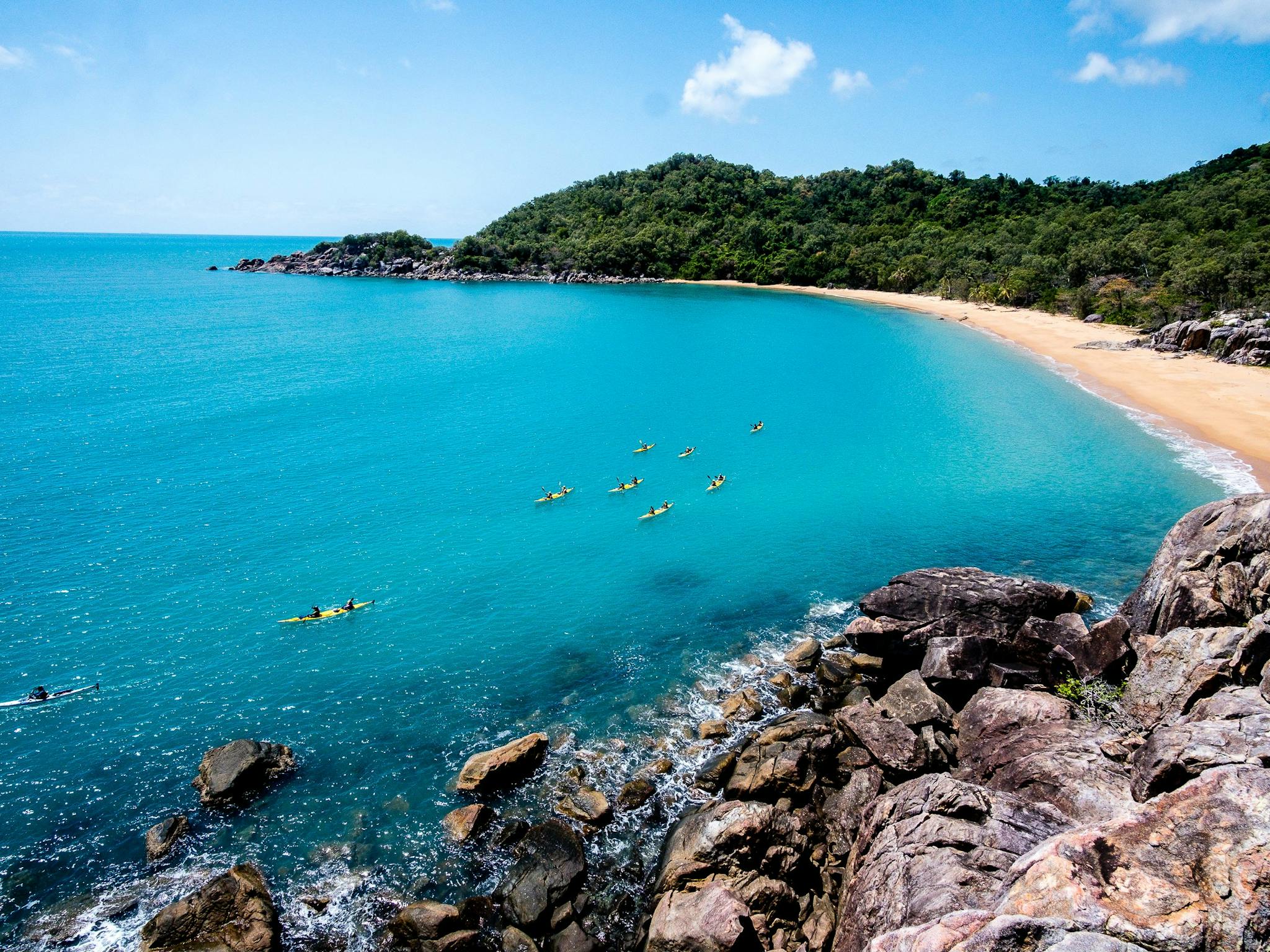 Sea Kayakers paddle across bright, clear blue water in front of a small bay flanked by rainforest Sea Kayakers paddle across bright, clear blue water in front of a small bay flanked by rainforest