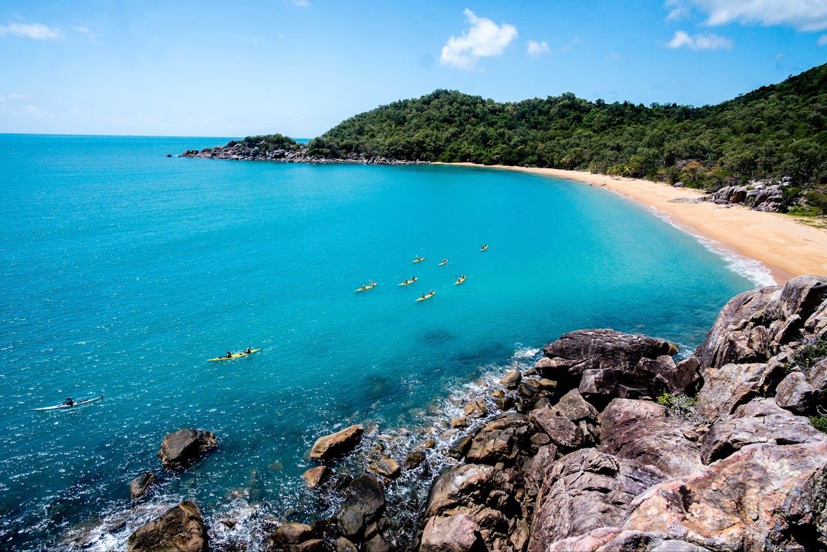 Sea Kayakers paddle across bright, clear blue water in front of a small bay flanked by rainforest