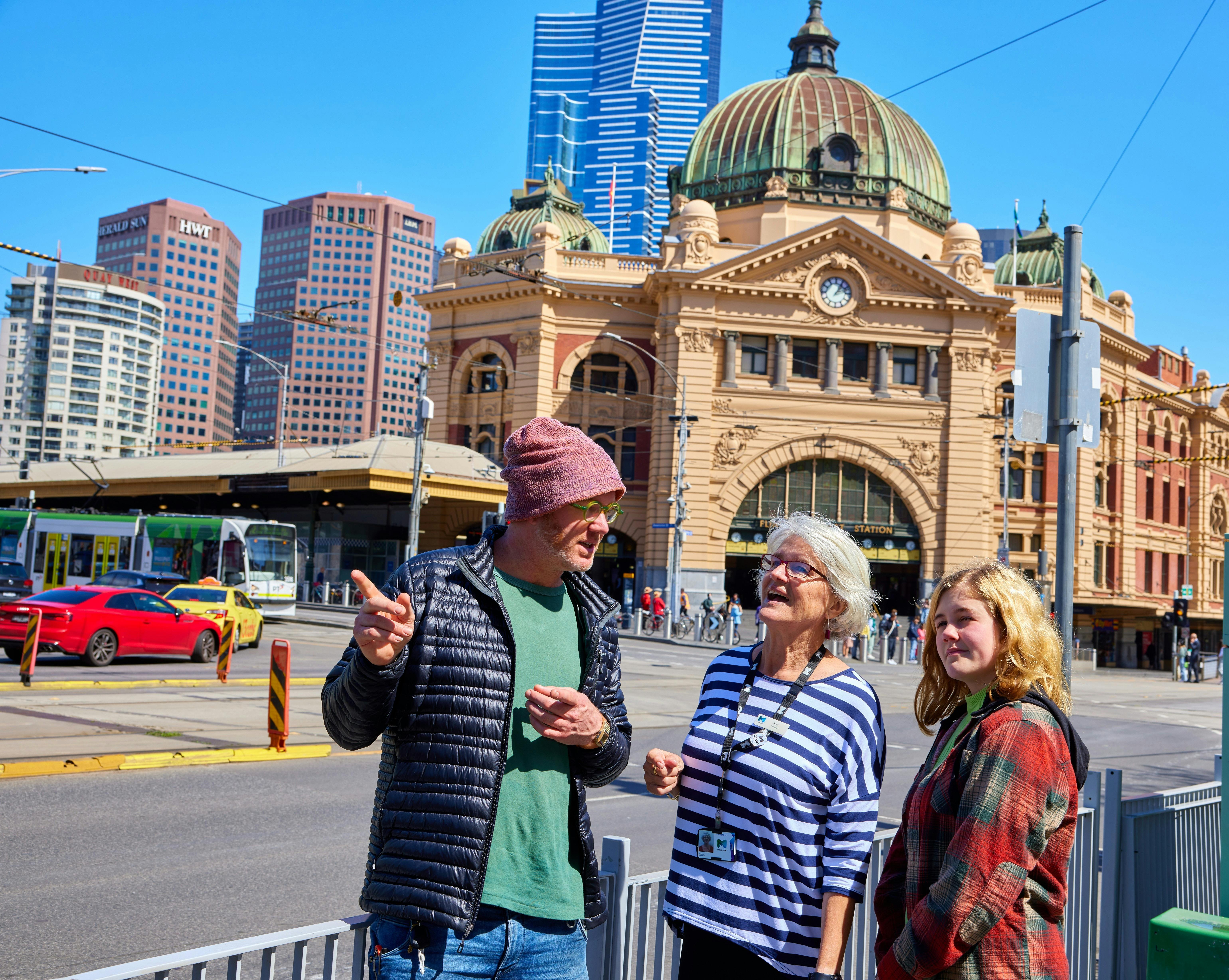 people looking in a direction being pointed by a third person with Flinders Street Station behind
