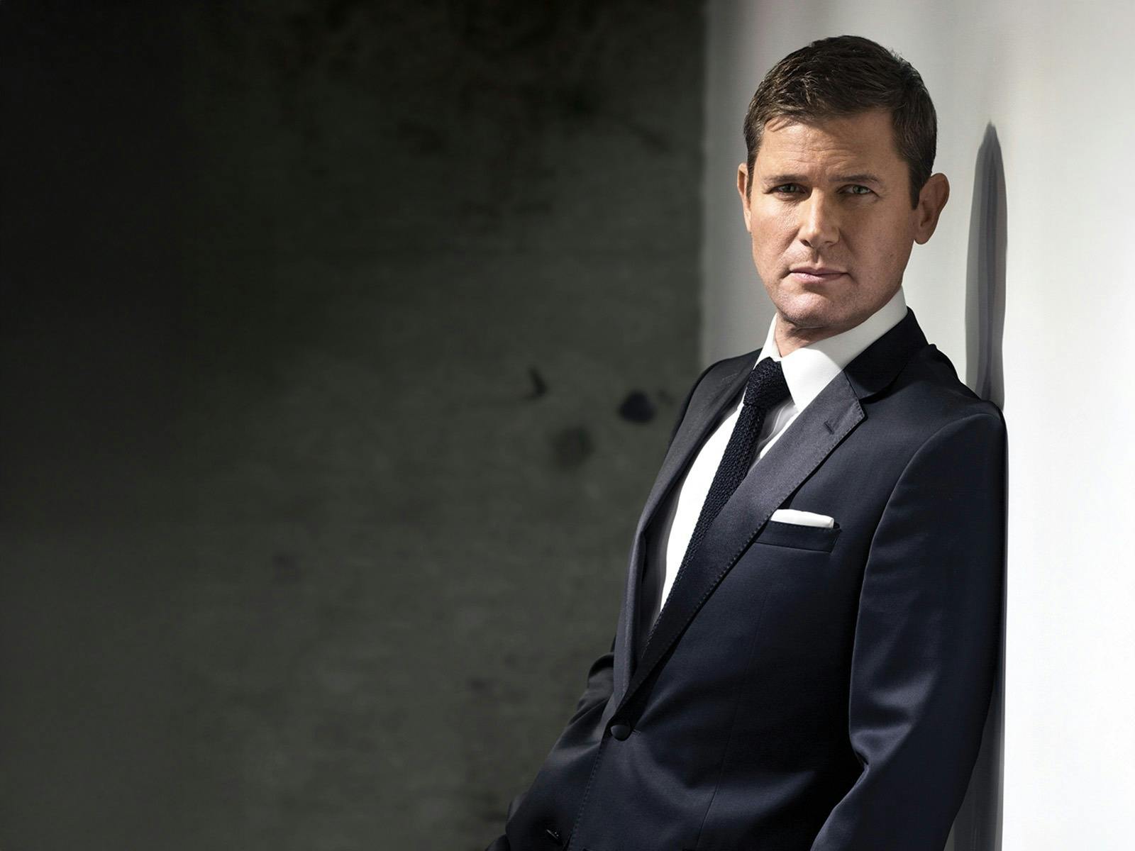 Man wearing a black suit leaning on a wall