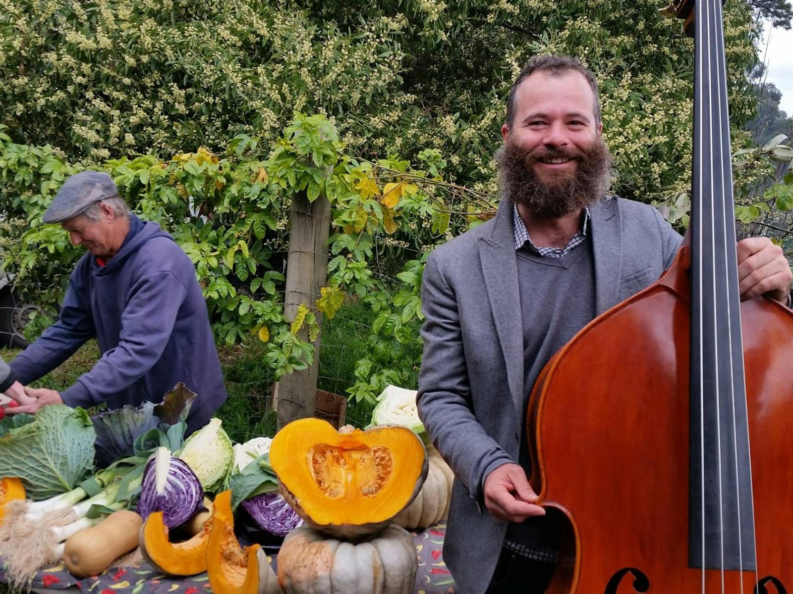 A person stands with a double bass in front of a table with farm fresh produce on it.