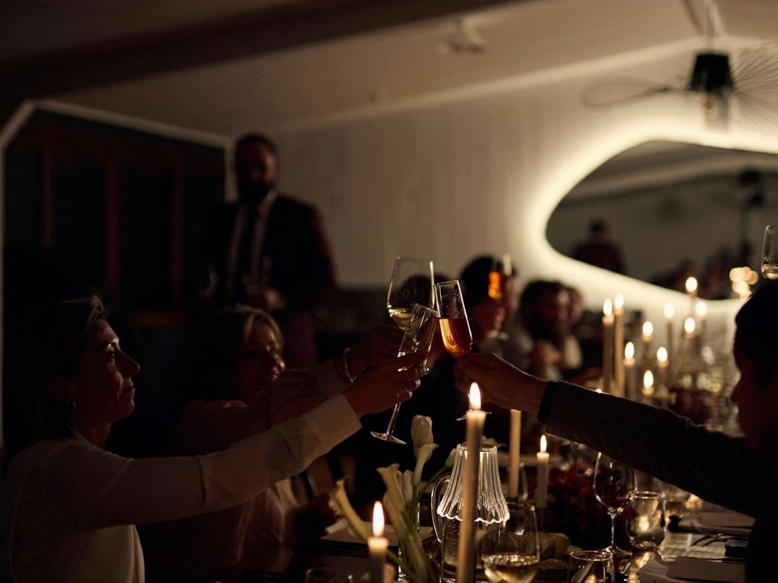 Guest gathered at a long table and clink glasses over dinner with candles and ambient lighting