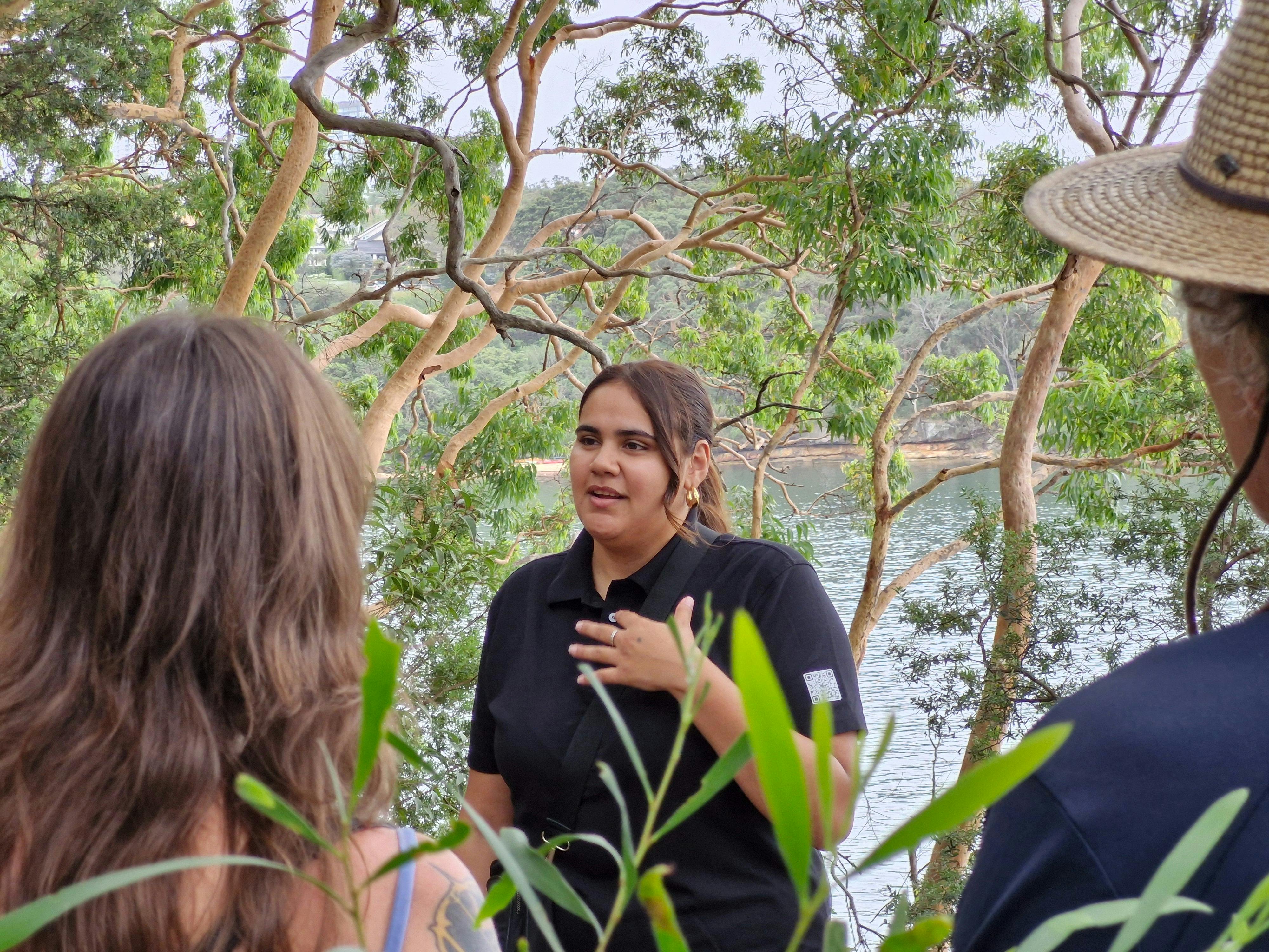 Junge Aborigine-Reiseführerin mit den Rücken von zwei Gästen, hinter der Reiseleiterin sind Eukalyptusbäume und Wasser zu sehen.