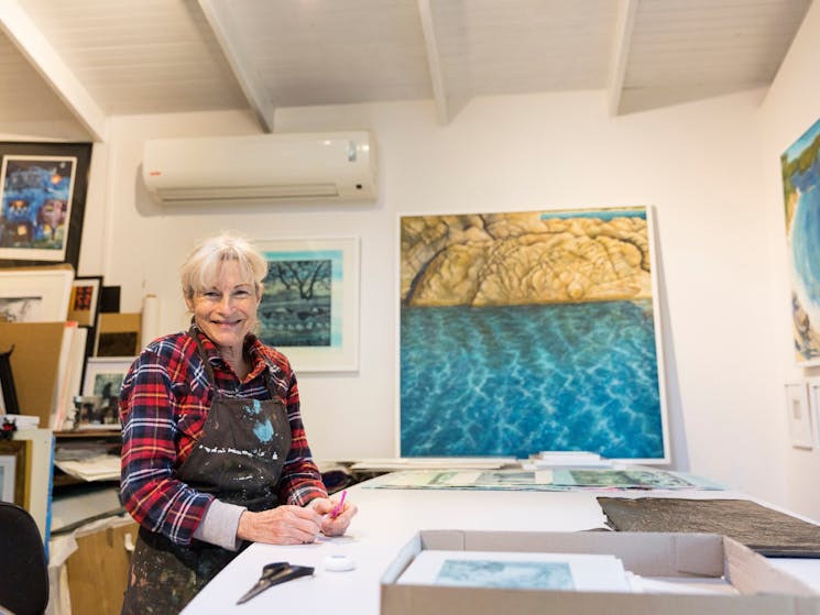 A white room with framed ocean paintings on the walls. The artist stands at her desk smiling at the