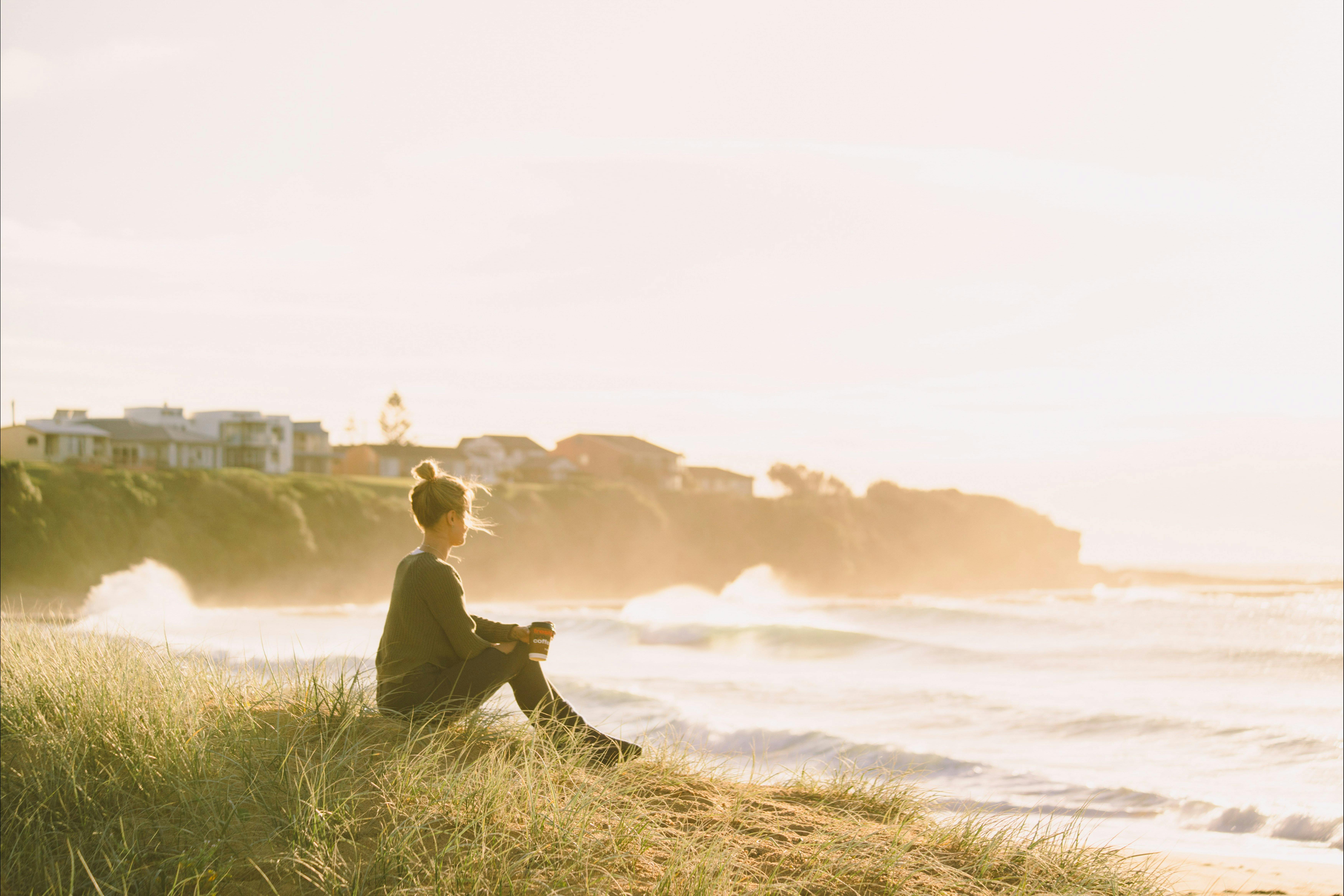 Woman sitting on sand dunes holding her coffee and watching the waves roll in