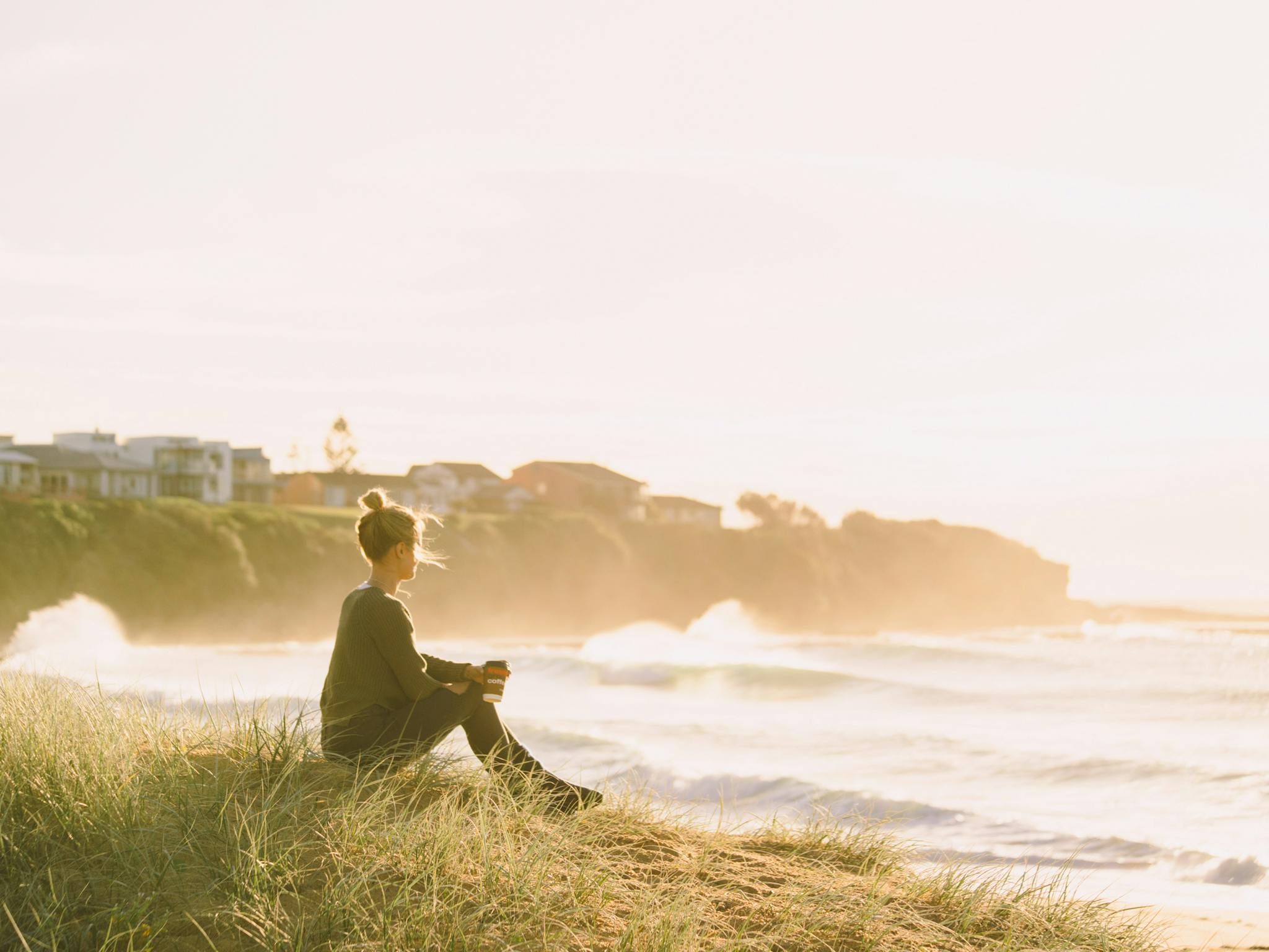 Woman sitting on sand dunes holding her coffee and watching the waves roll in
