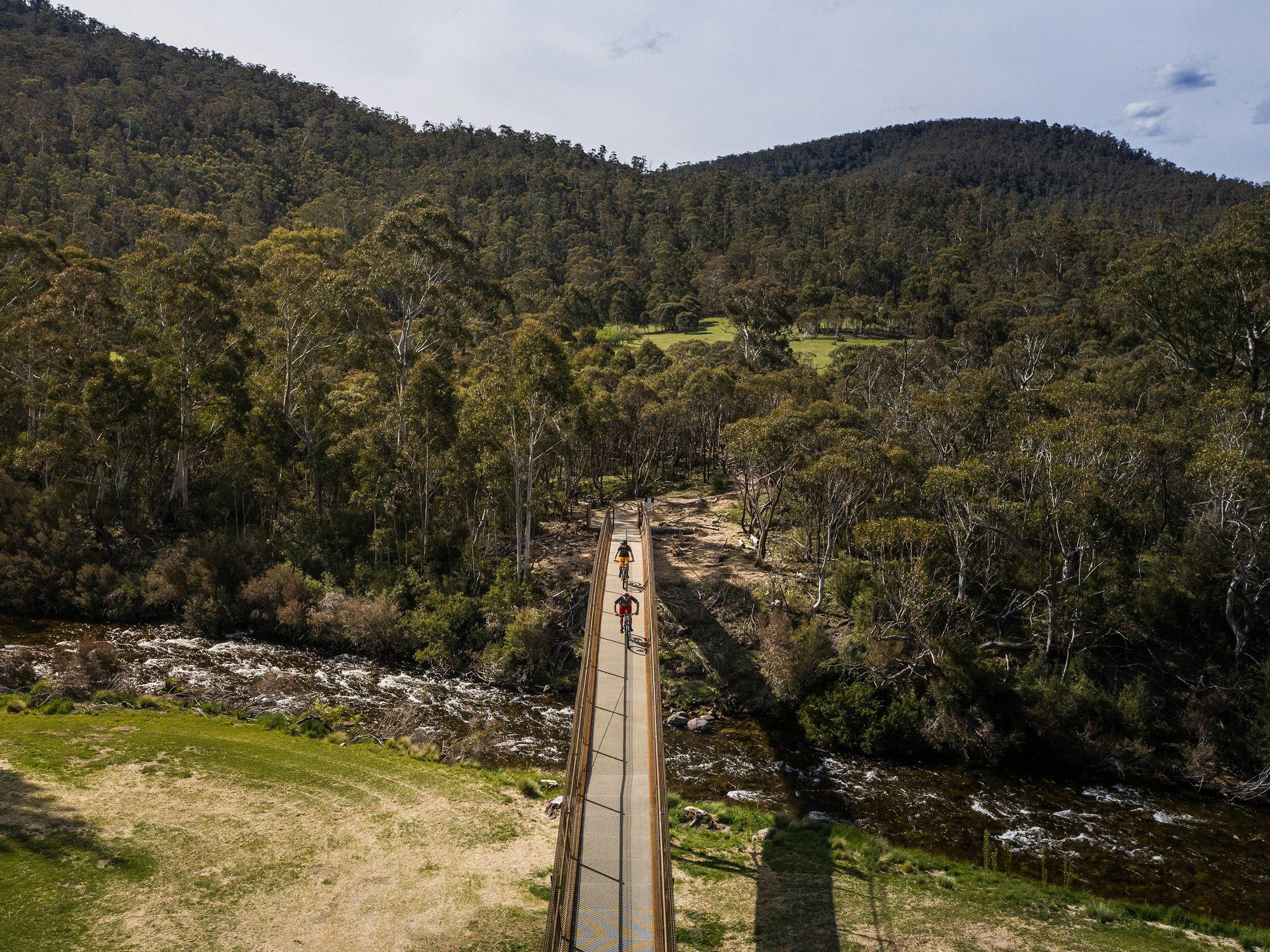 Aerial view of two bike riders crossing a bridge on Thredbo Valley track to Gaden Trout Hatchery
