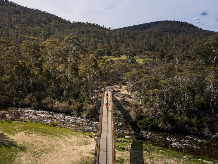 Aerial view of two bike riders crossing a bridge on Thredbo Valley track to Gaden Trout Hatchery