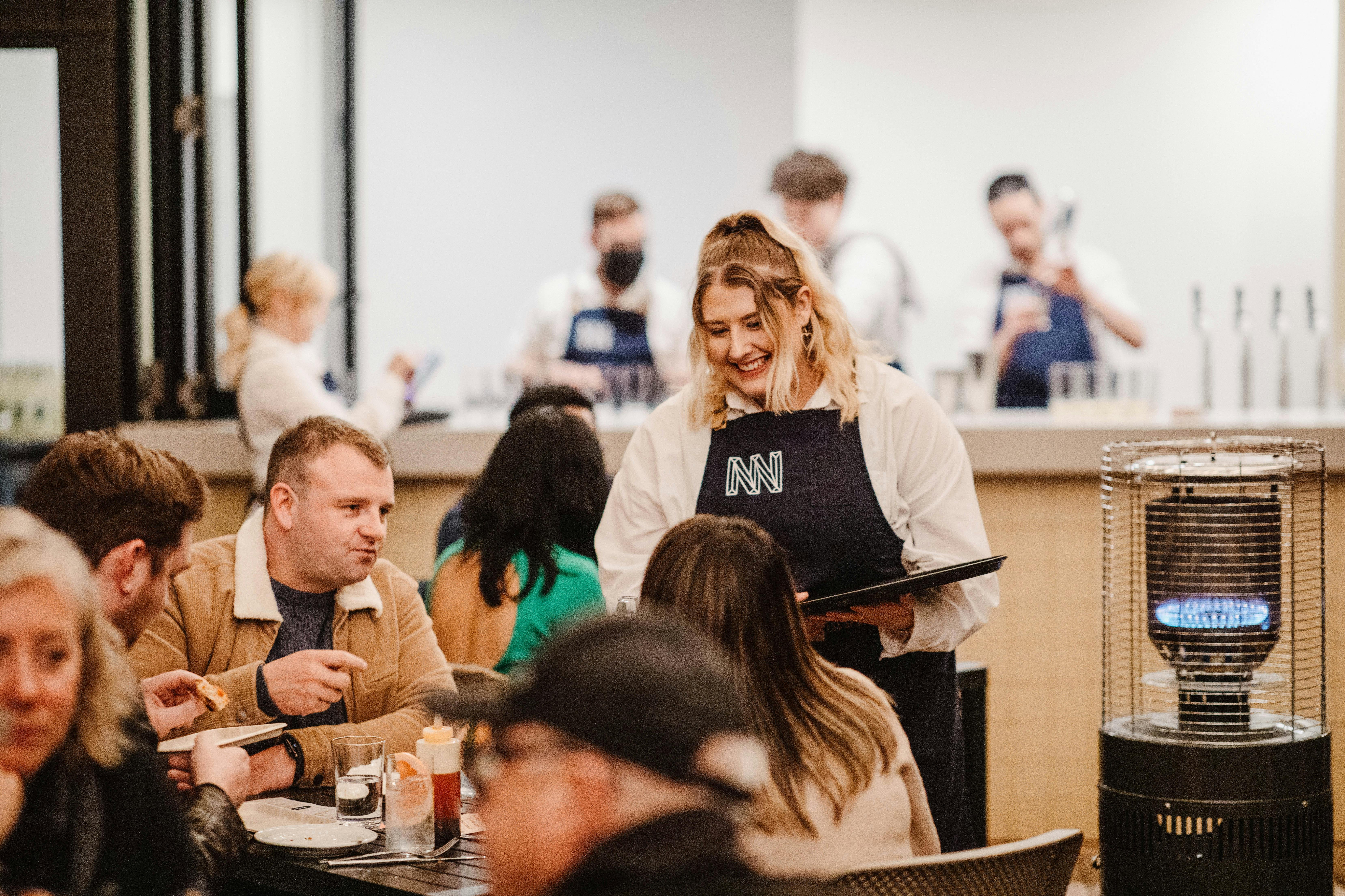 a waitress smiles at a table of happy guests enjoying some pizzas and gin and tonics