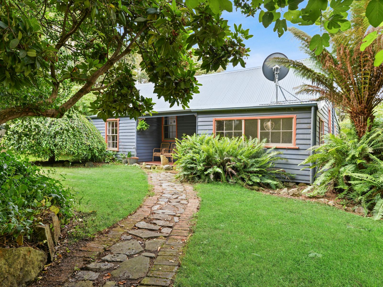 A wooden 1900s miners cottage painted smoky blue with a crooked stone path amid a lush green garden