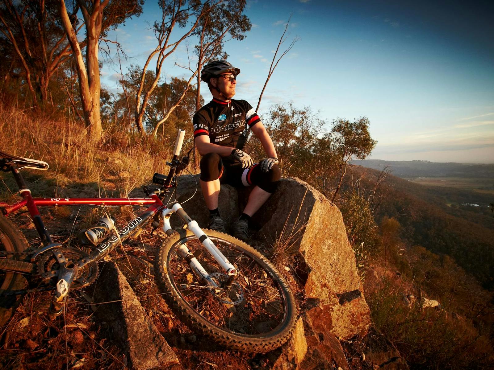 Cyclist sitting on rock, mountain bike lying on ground,  gum trees, lookout, distant hills