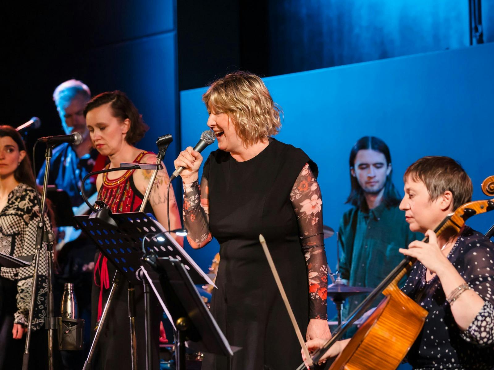 A female singer performing into a microphone on stage with a live band and cellist.