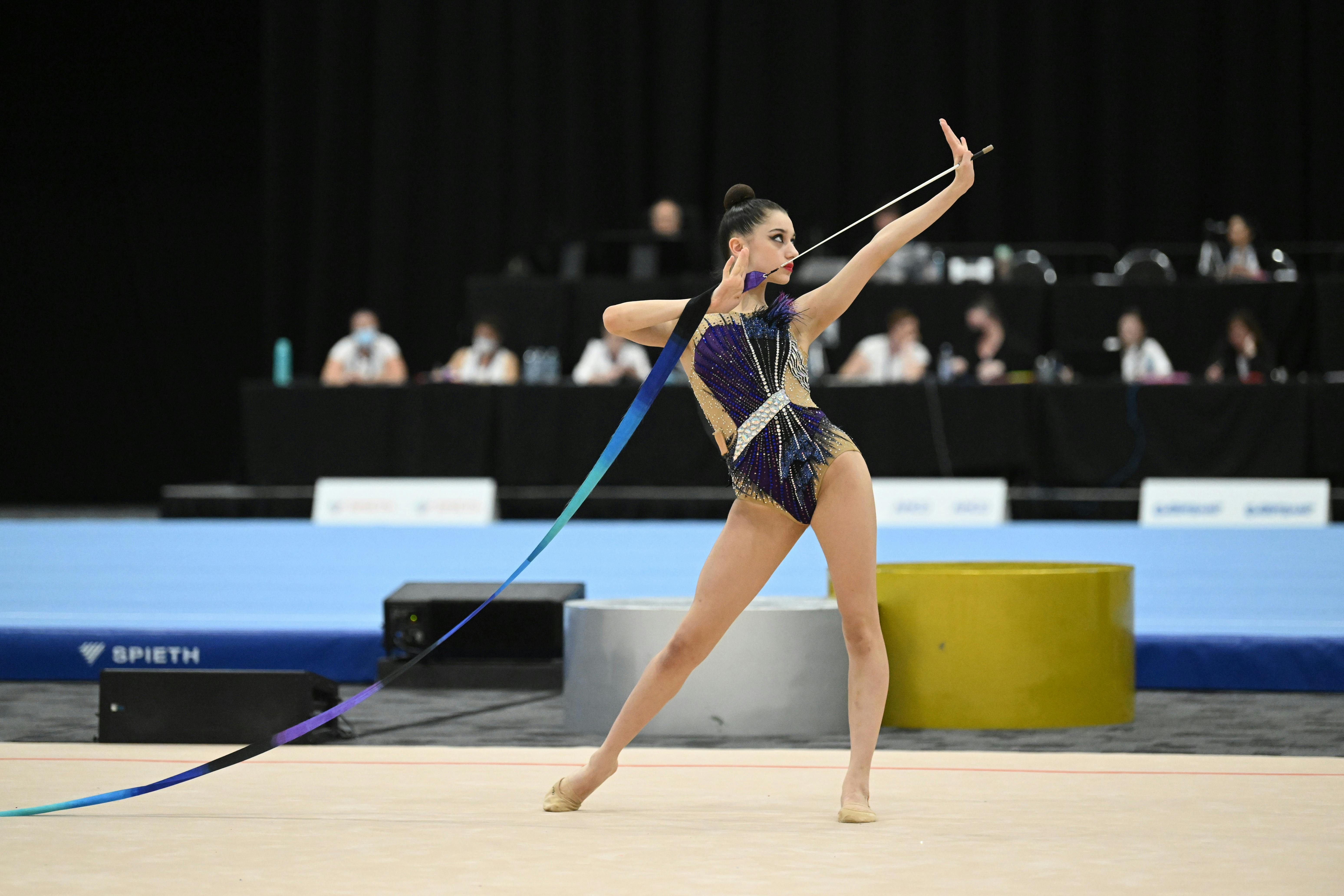 Gymnast in black leotard poses with arm outstretched and blue ribbon in hand