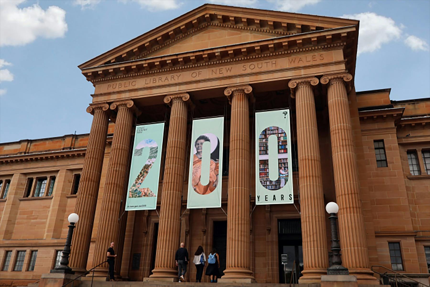 A grand sandstone building with banners between the ionic columns that spell out '200'
