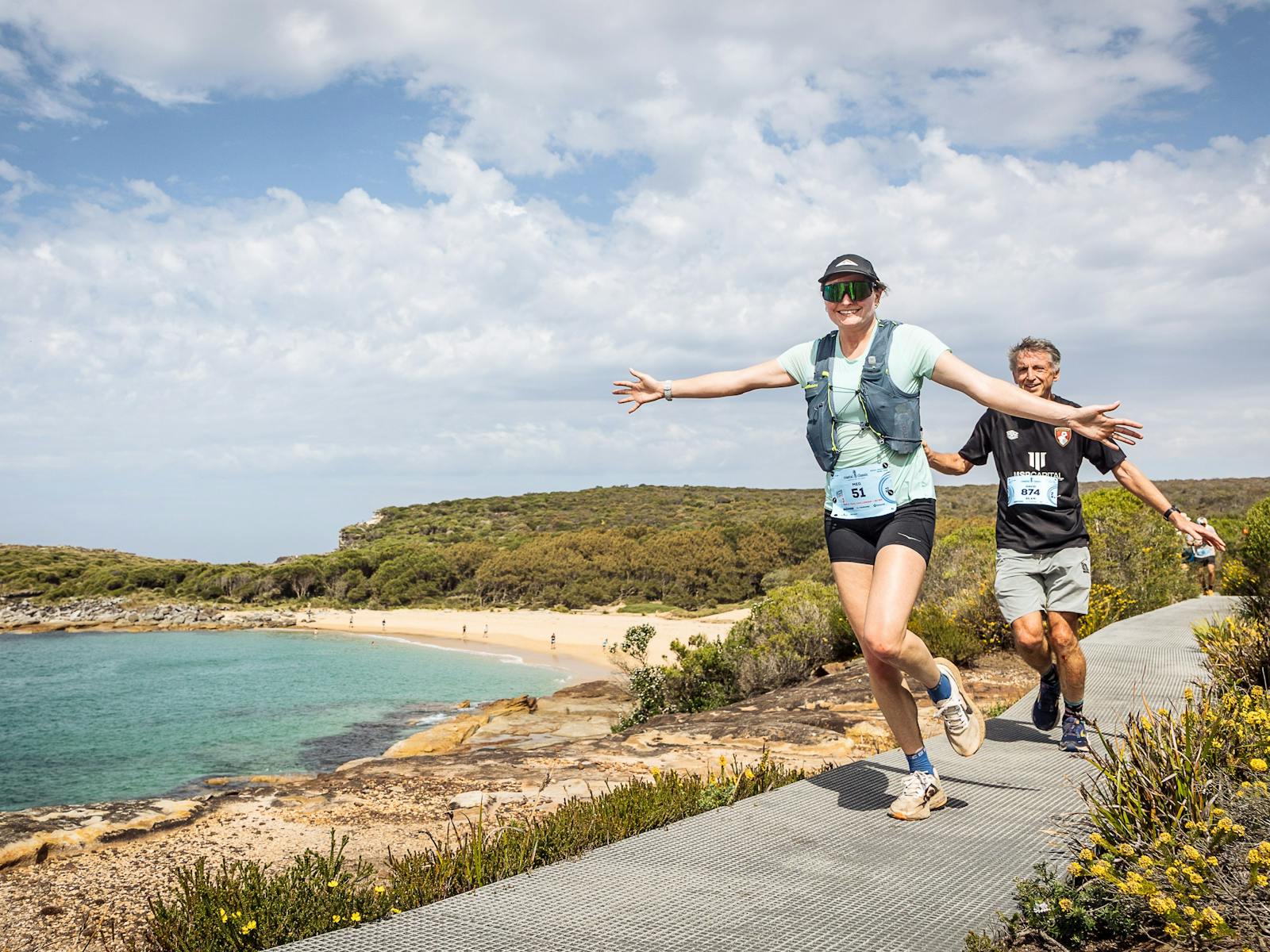 Two runners smiling with their arms outstretched on coastal boardwalk.