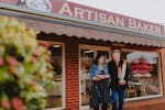 A couple exiting the Artisan Baker looking happy. The man is holding a baguette.