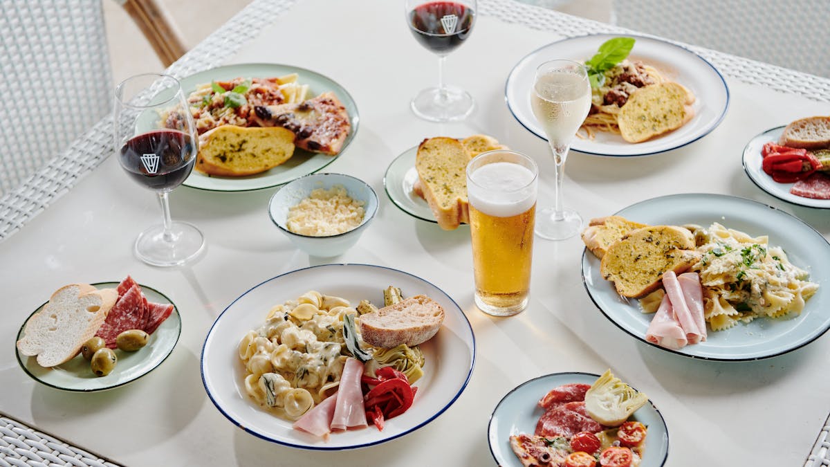 Pizza and pasta dishes on a marble table with a beer and two glasses of red wine