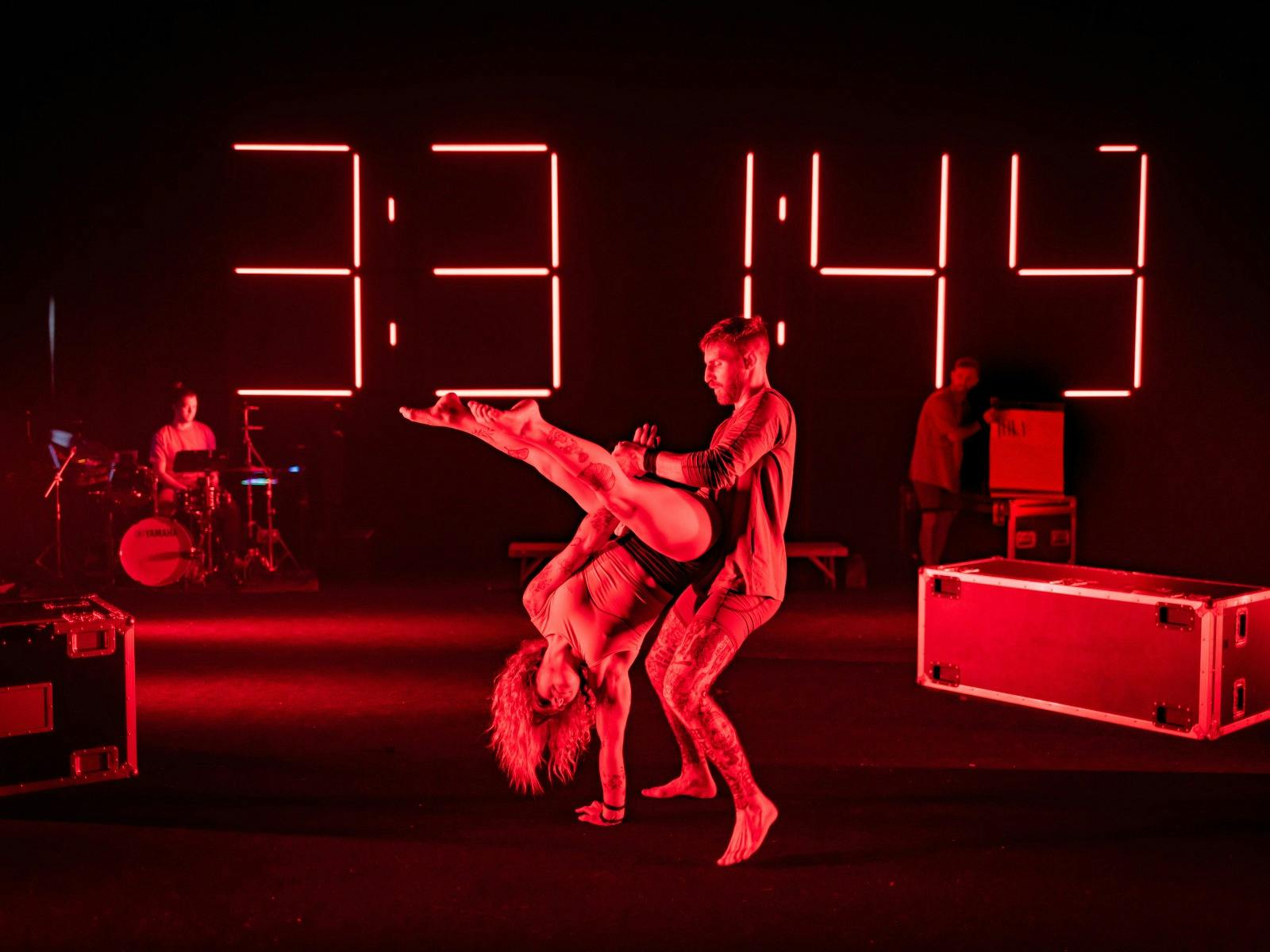 A group of acrobats performing stunts lit by coloured lighting with a neon clock in the background.