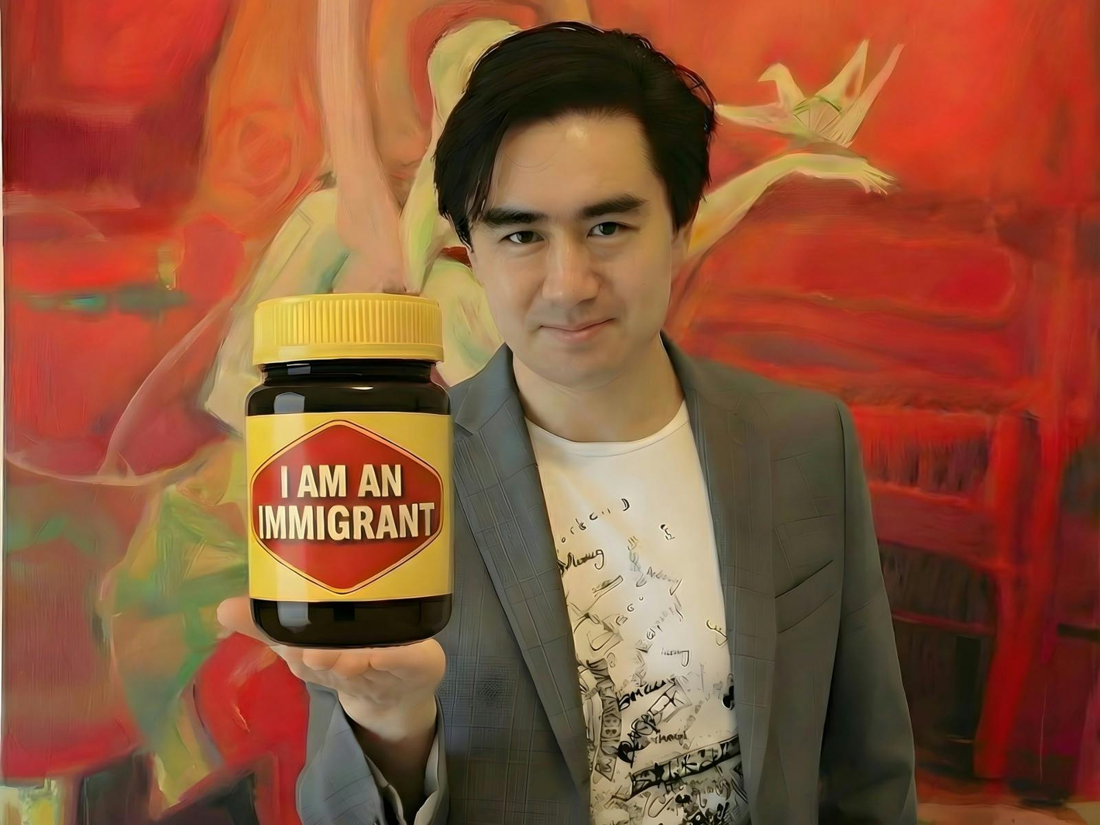 A smiling person in a gray blazer holds a vegemite jar labeled “I AM AN IMMIGRANT” .