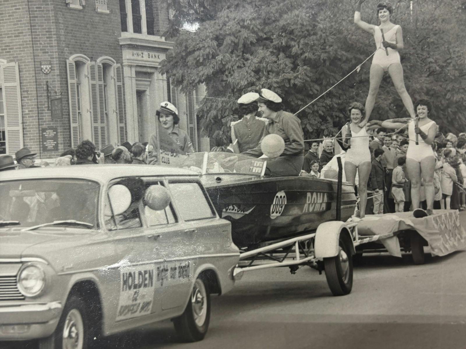 A parade, a car pulls a boat, complete with 3 women forming a standing pyramid