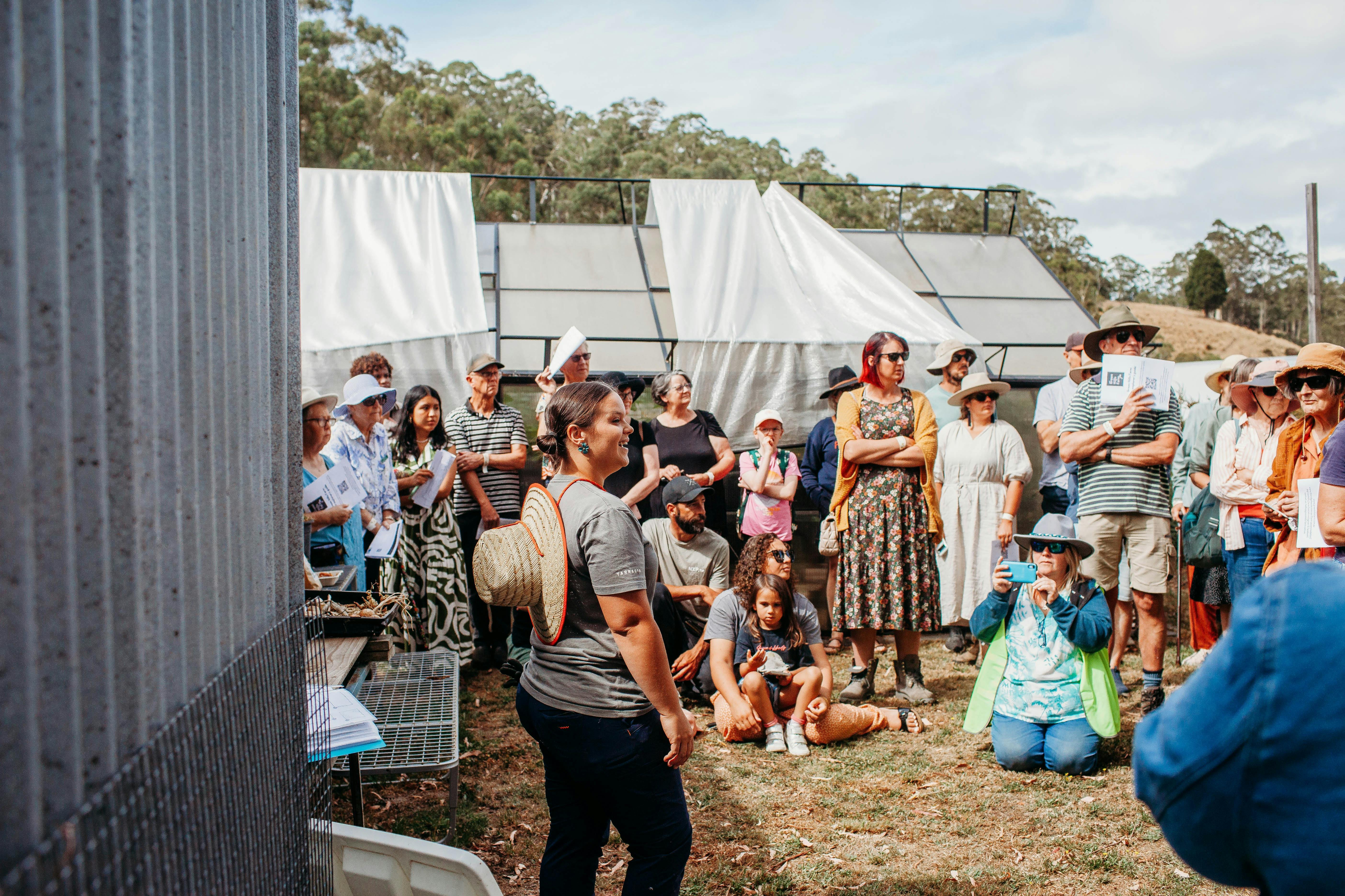 A lady with a t-shirt and a straw hat is facing a group of people listening to her talk.