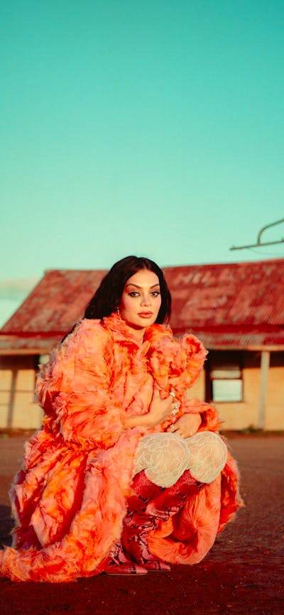 Gamilaraay musician Thelma Plum, wearing a coral-coloured feather coat, kneels on Wilyakali Country.