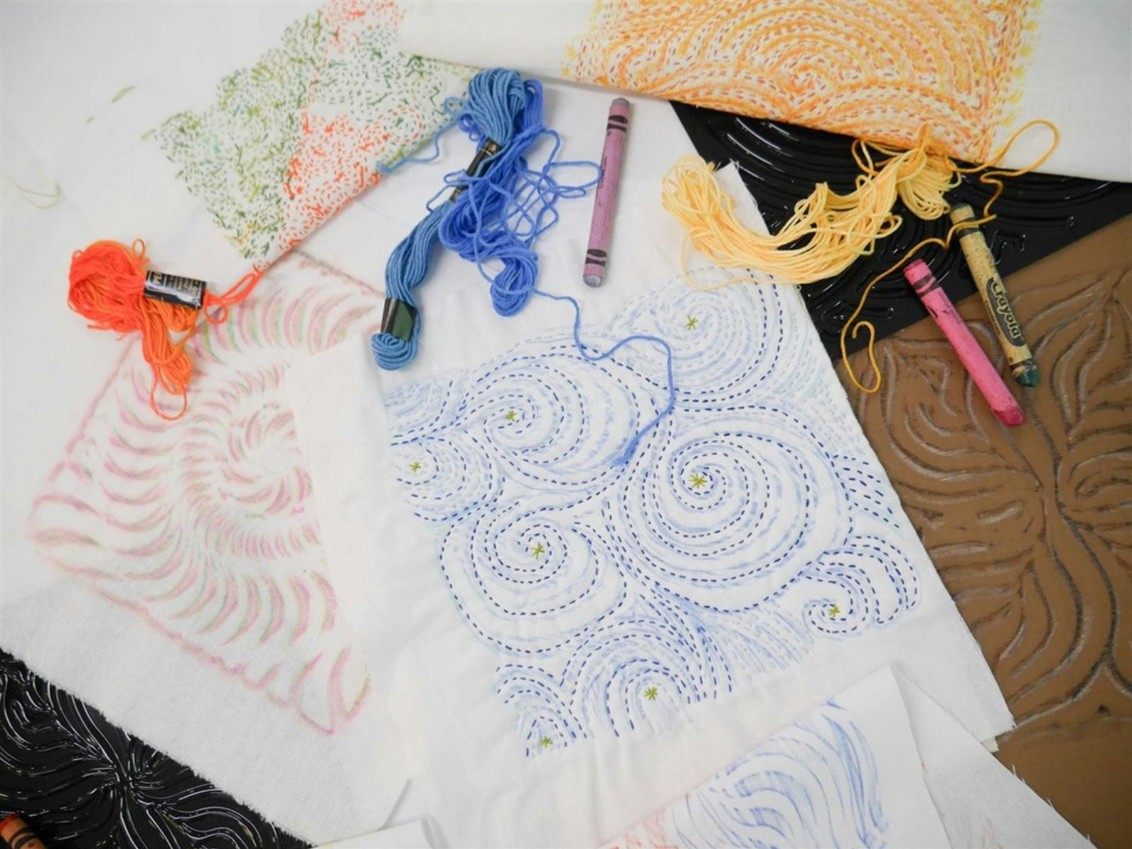 Flat lay photograph of embroidery piece, string, crayons in blue pink and yellow colours.