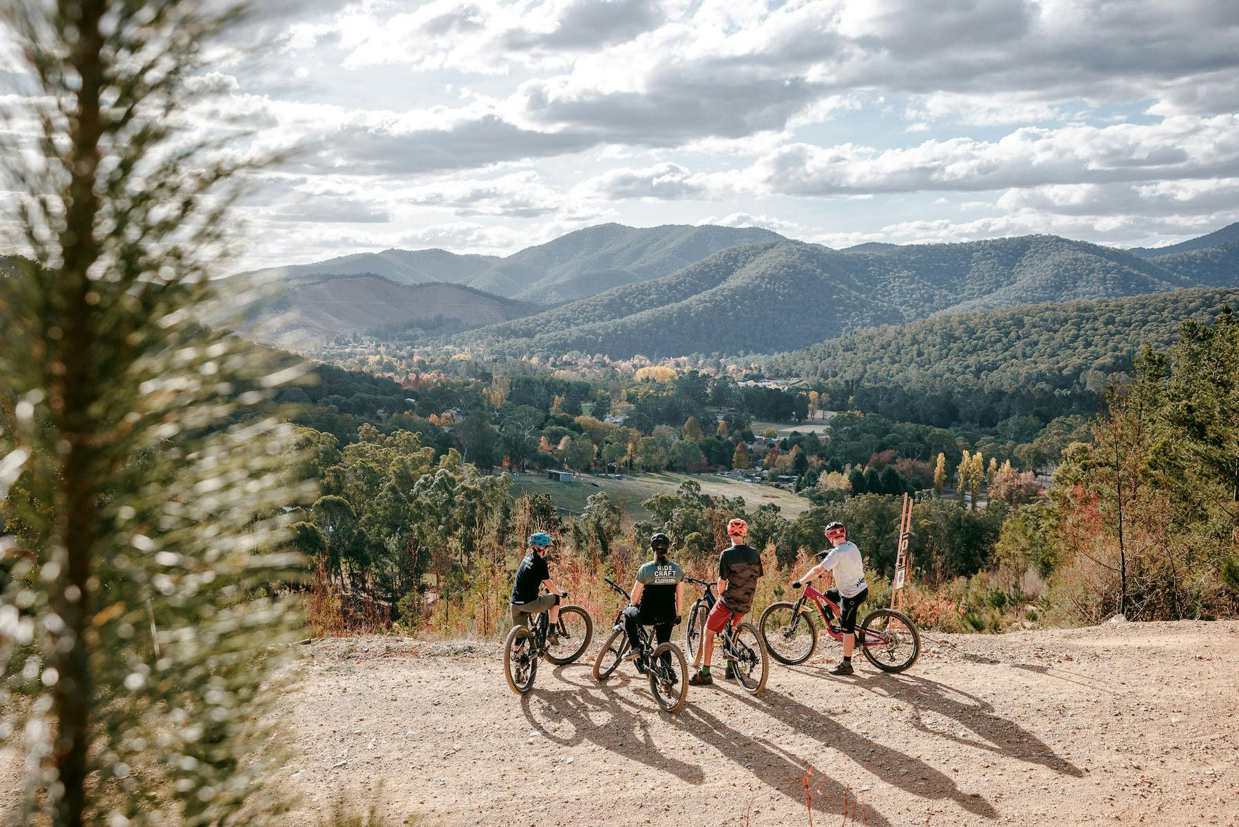 Four riders stand with their backs to the camera overlooking a valley with mountain views