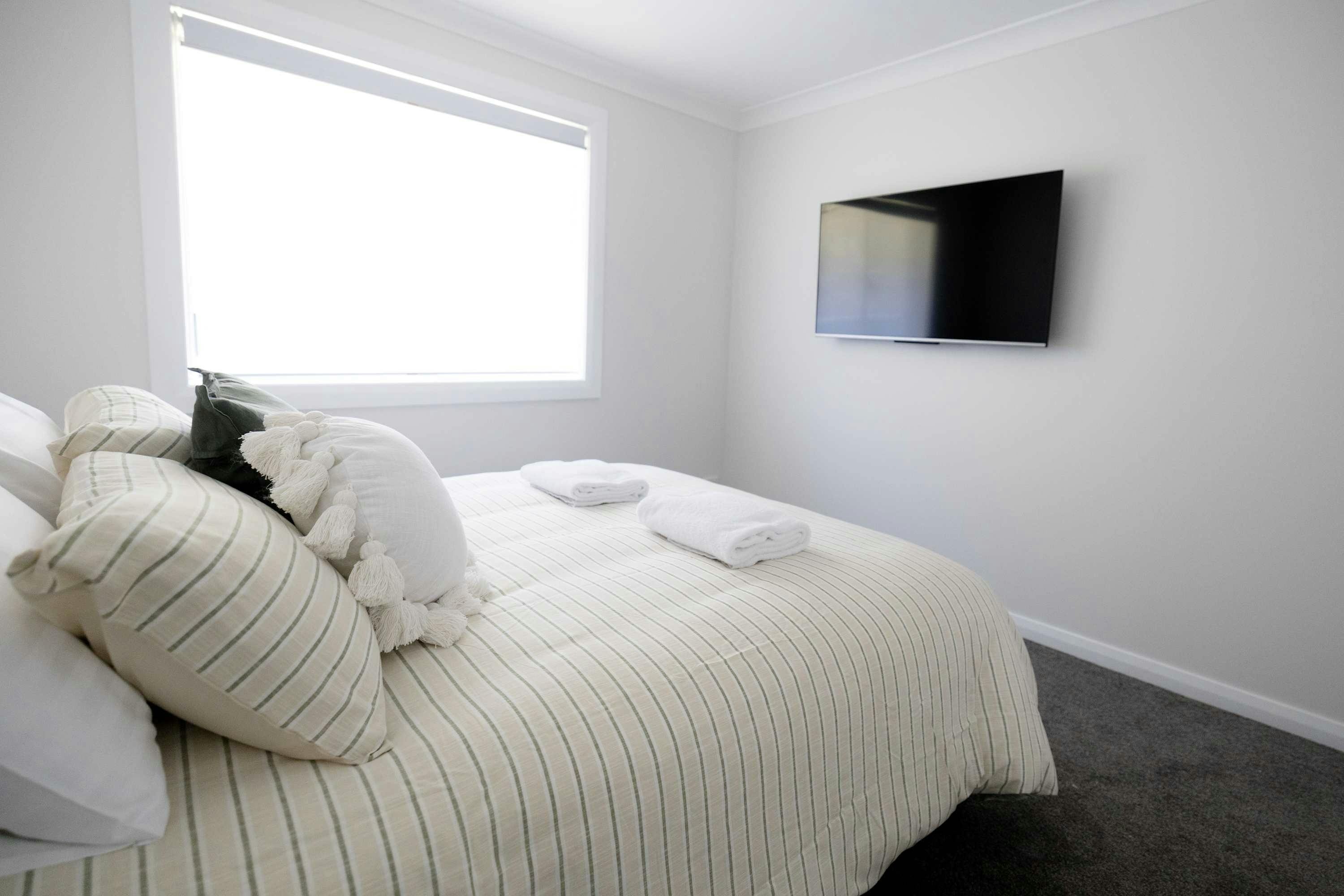 Queen bed with neutral bed sheets, large window for natural light and a tv mounted to the wall
