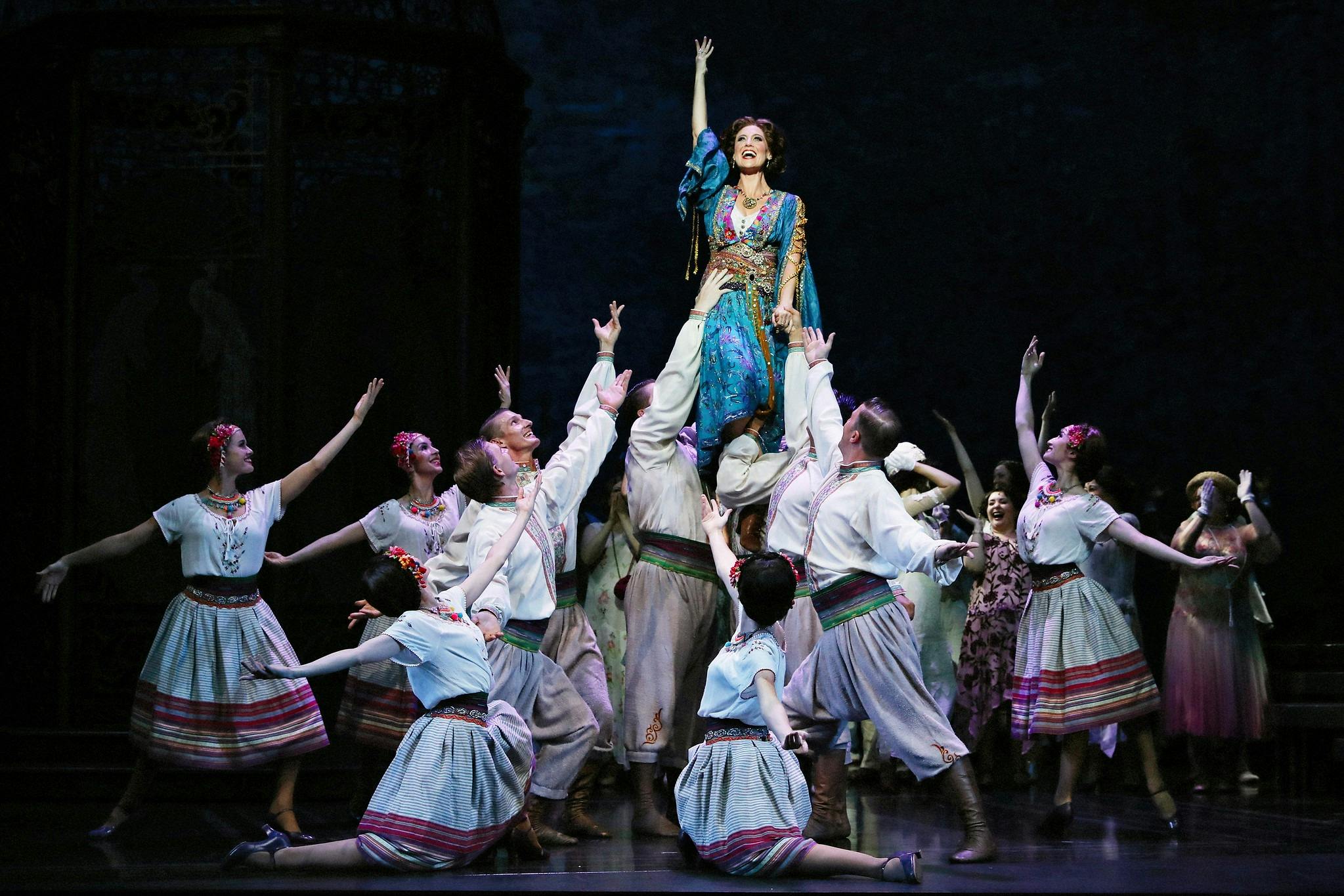 Woman in blue gown is held high by dancers in peasant costumes who surround her in a big song finish