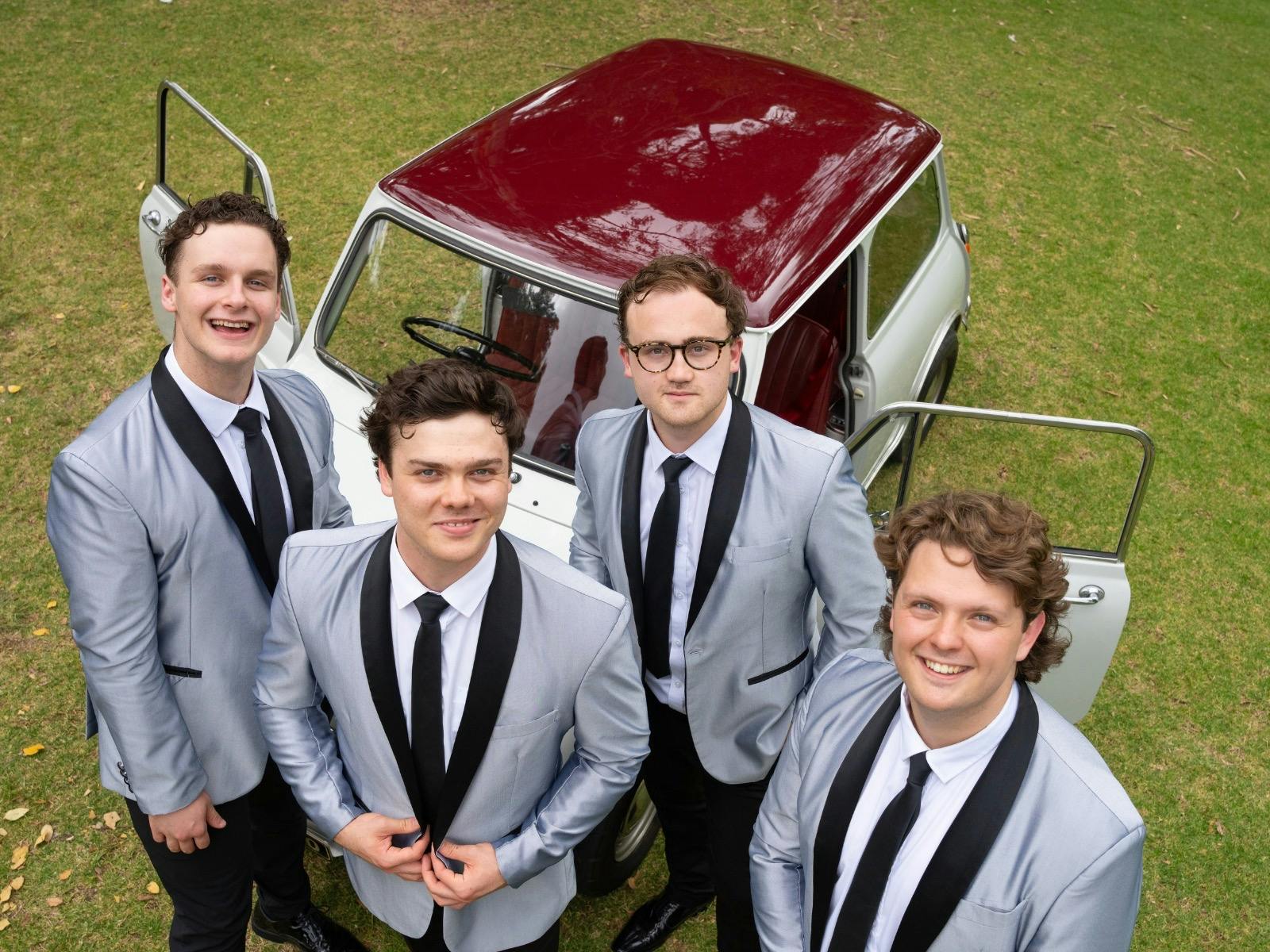 The band stands outdoors near a vintage car, smiling in matching silver jackets and black ties.