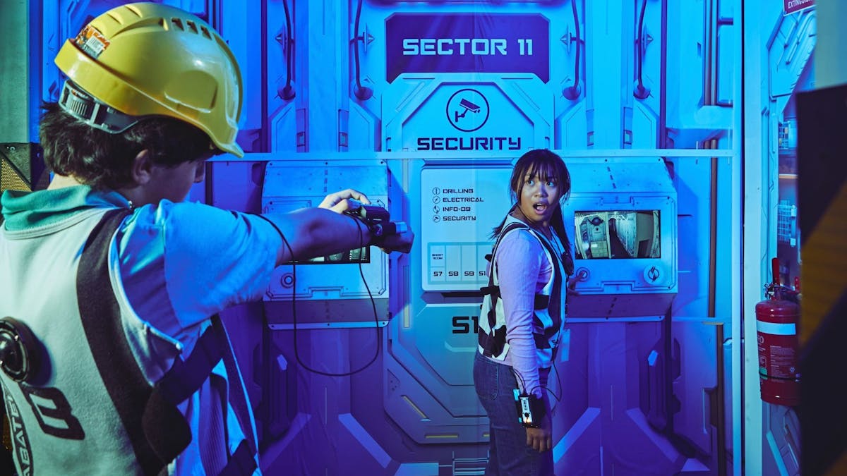 A boy points a wrist device at a shocked girl inside a blue-lit sci-fi room, Sector 11.