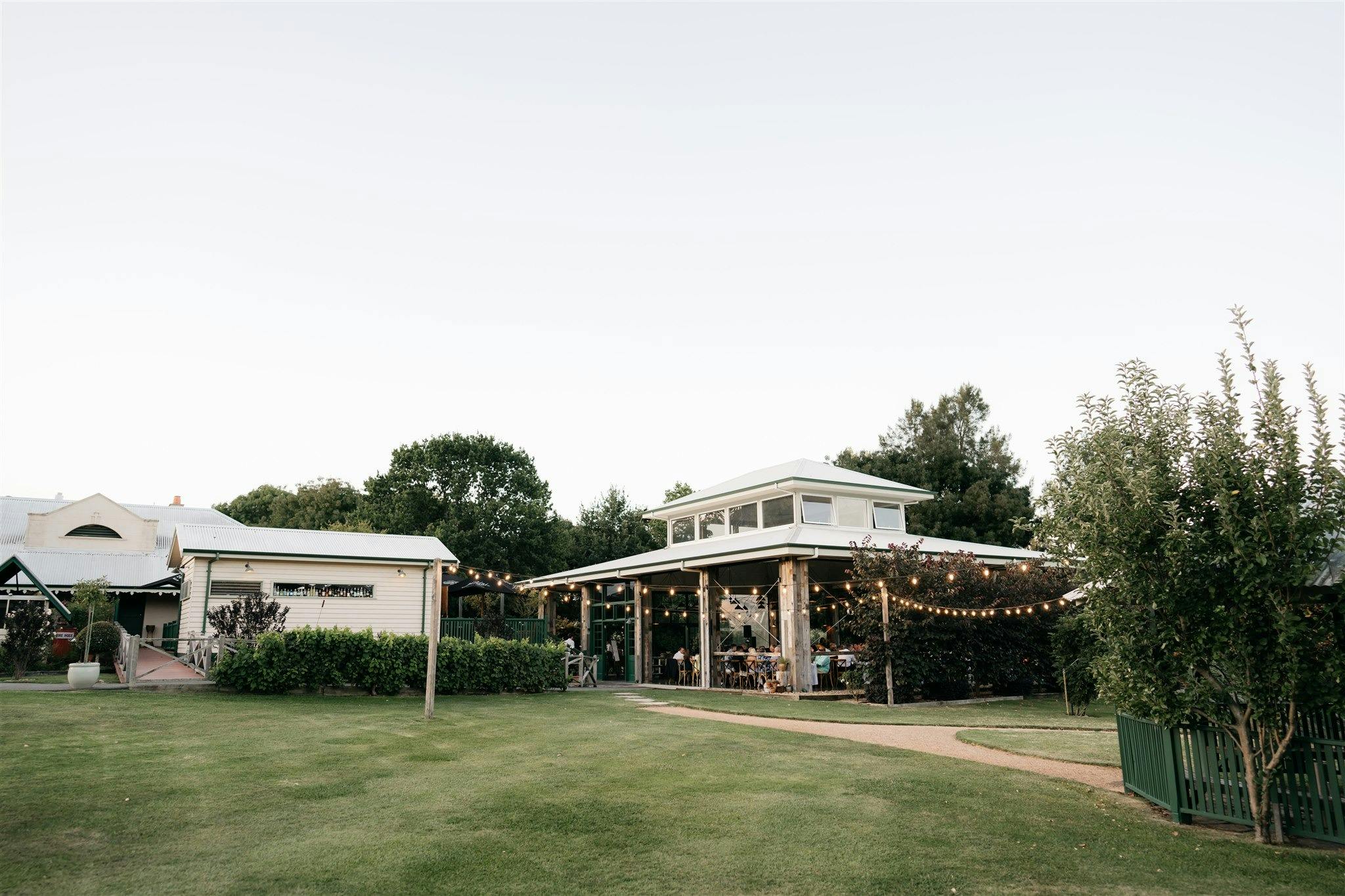 Outdoor venue with a pavilion, string lights, lush greenery, and guests dining in a warm, relaxed se