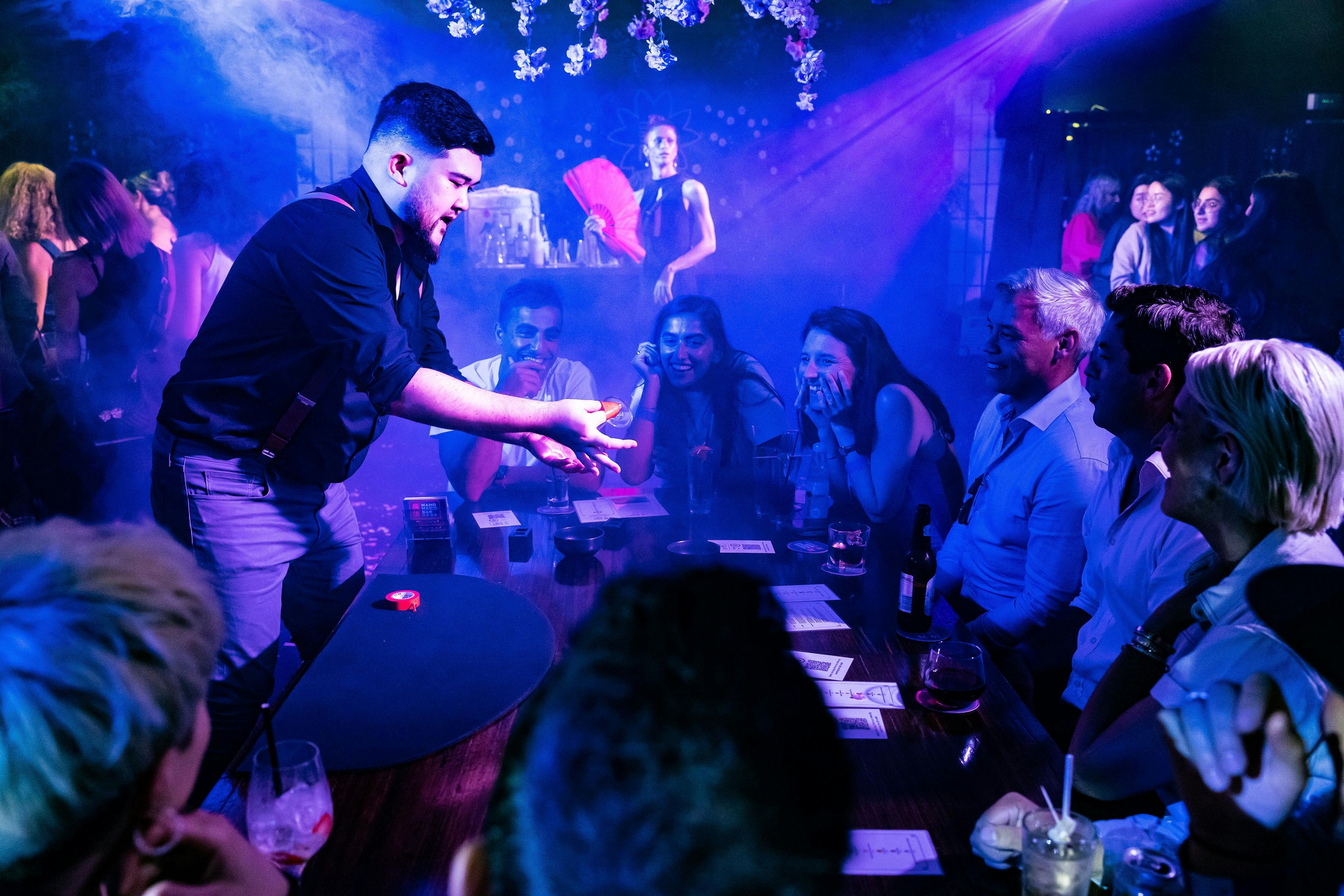 A table of people look on in wonder at a magician holding an object with their fingers taped.