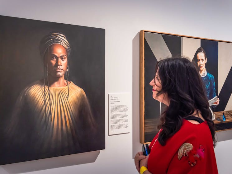 A woman in a red coat looks at a realistic portrait of a man in a turban and robes.