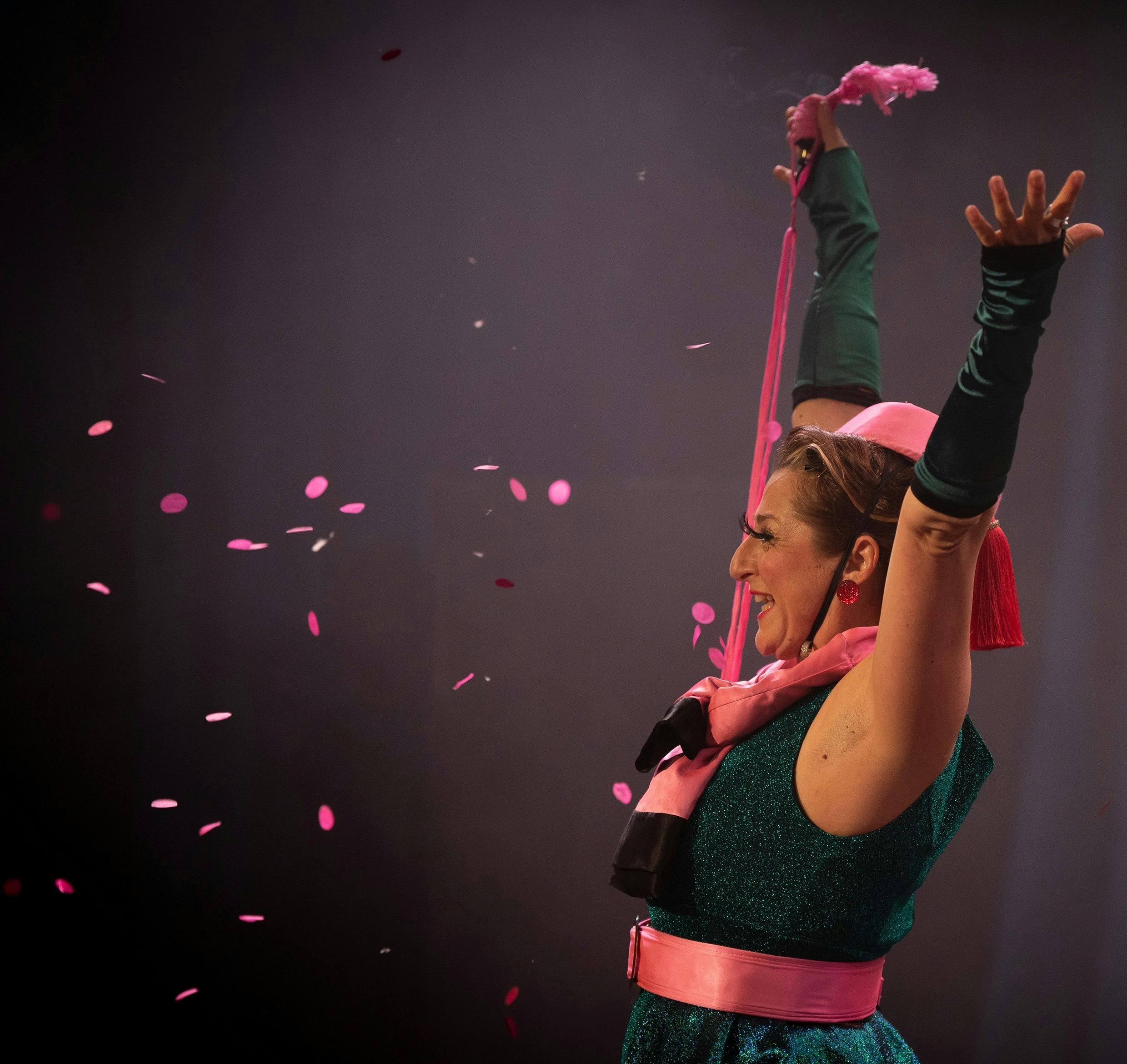 Glamorous woman in 1950s green wiggle dress, pink pillbox hat smiling with arms raised