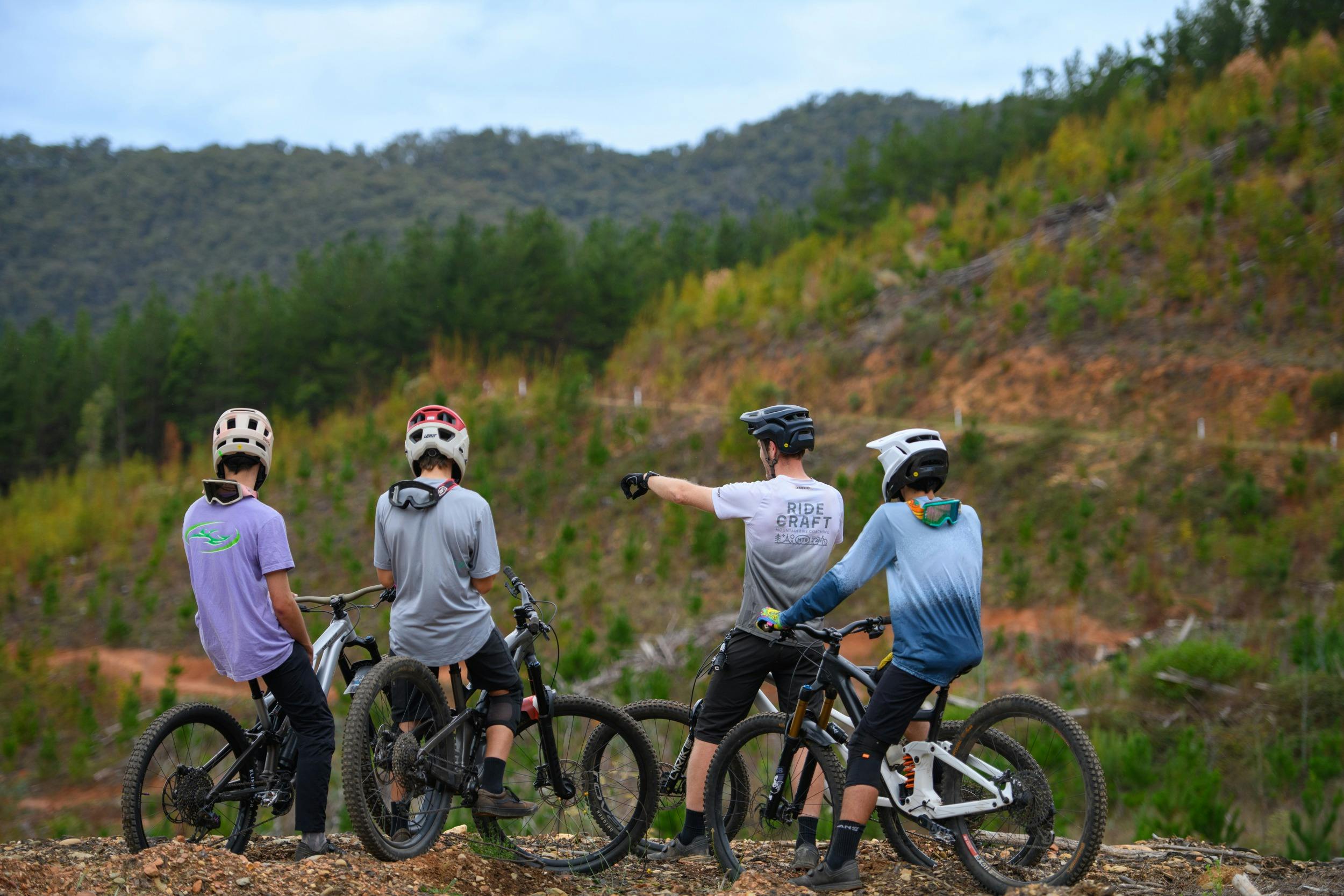 A group of bike riders sit on bikes with their backs to the camera, looking out over a pine forest.