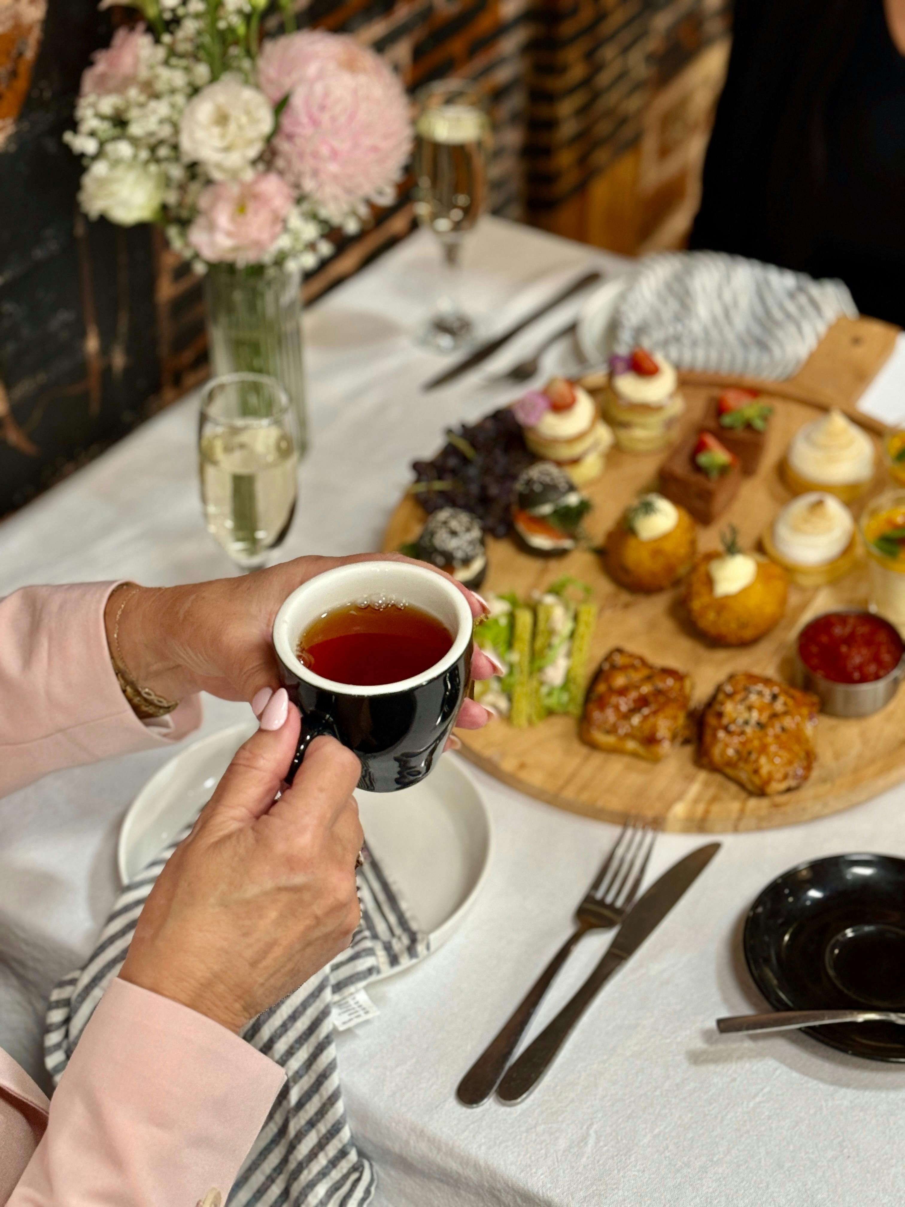 Holding a cup of tea over snacks at a table with white cloth, a platter of snacks and flowers