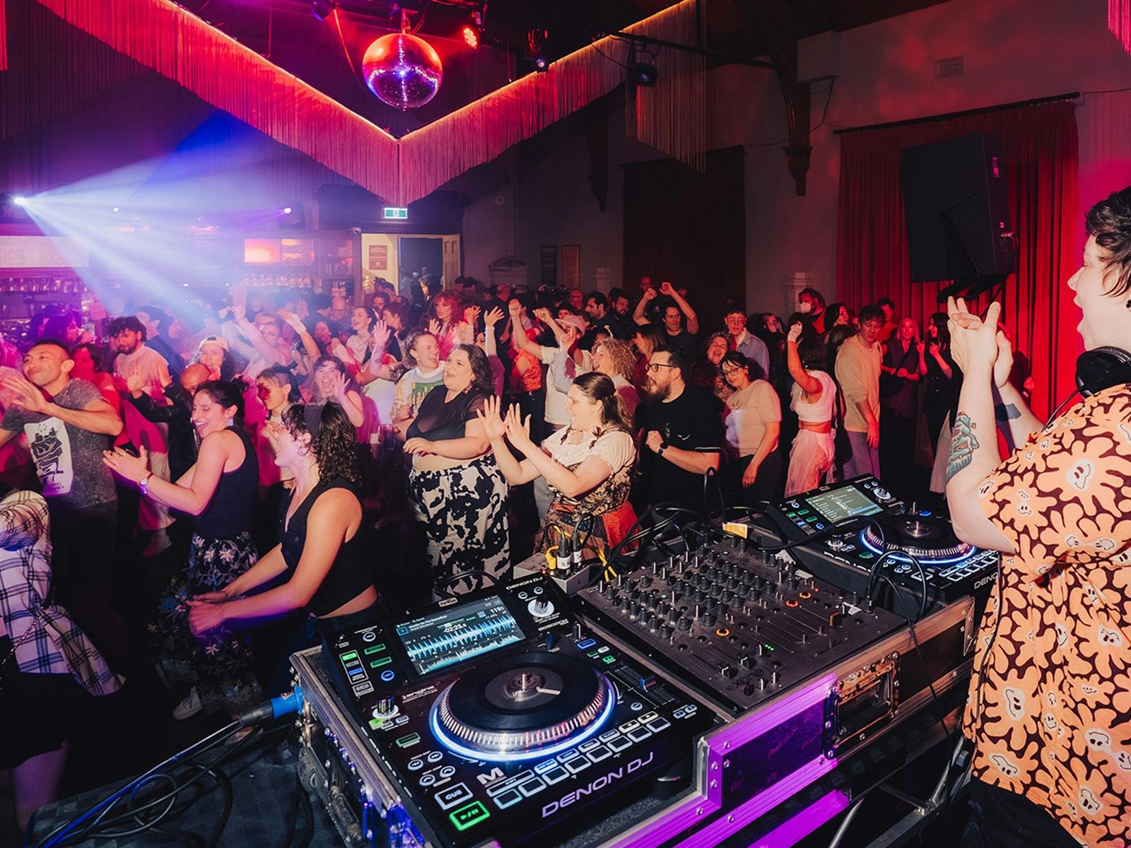A crowd dances in unison undr a disco ball while a DJ claps them on.