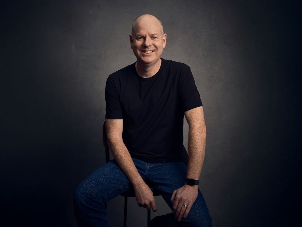 Comedian Tom Gleeson sits alone awkwardly smiling on a dark background