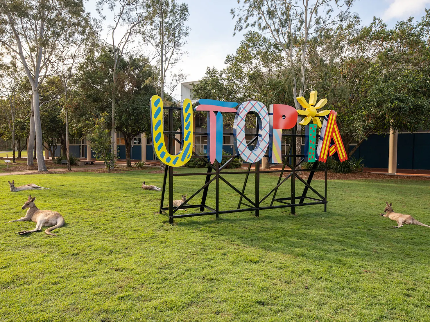 A colourful outdoor sign reads utopia with five kangaroos laying on the grass around it..