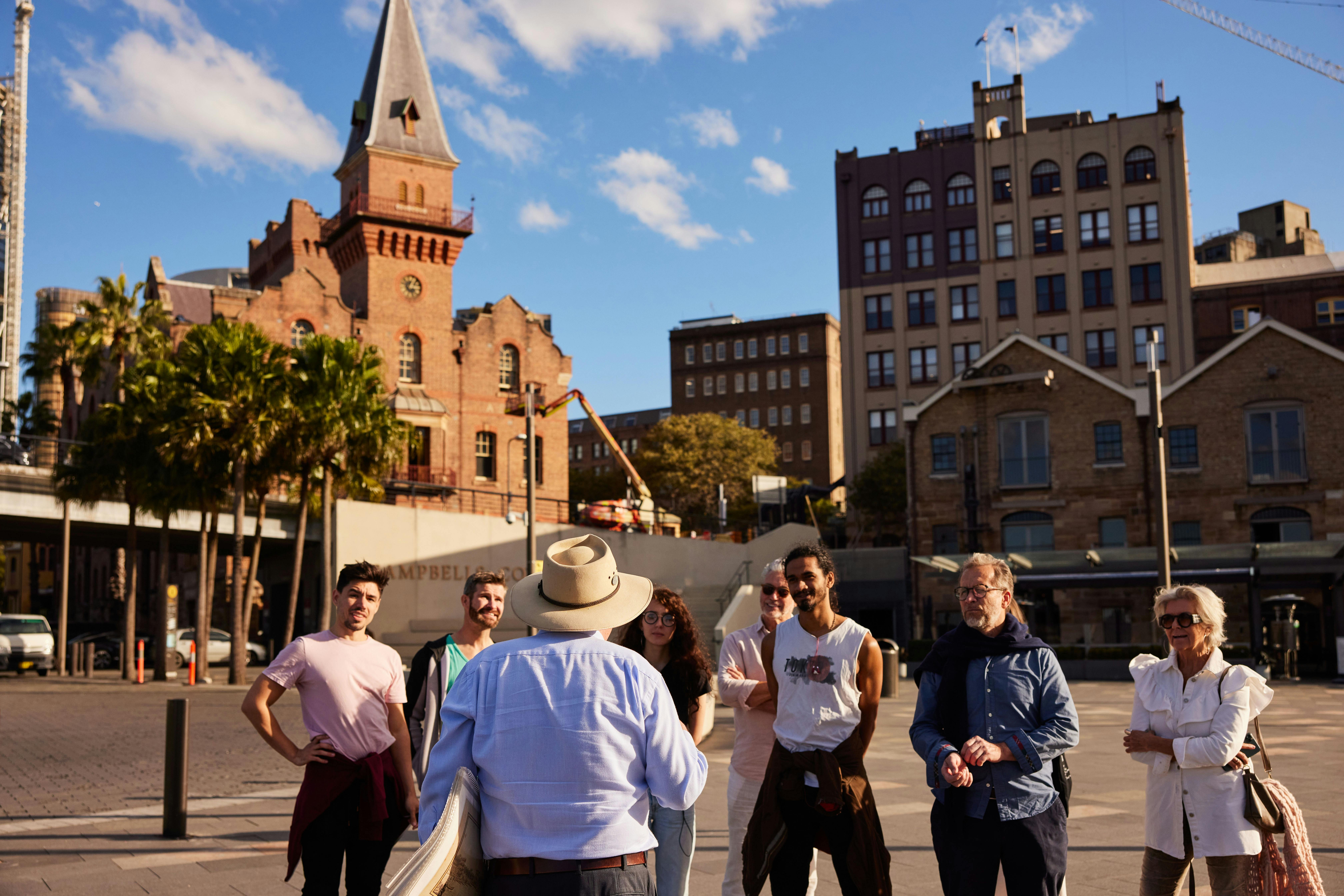 A walking tour guide speaks to a small group in The Rocks, Sydney, with historic brick buildings and