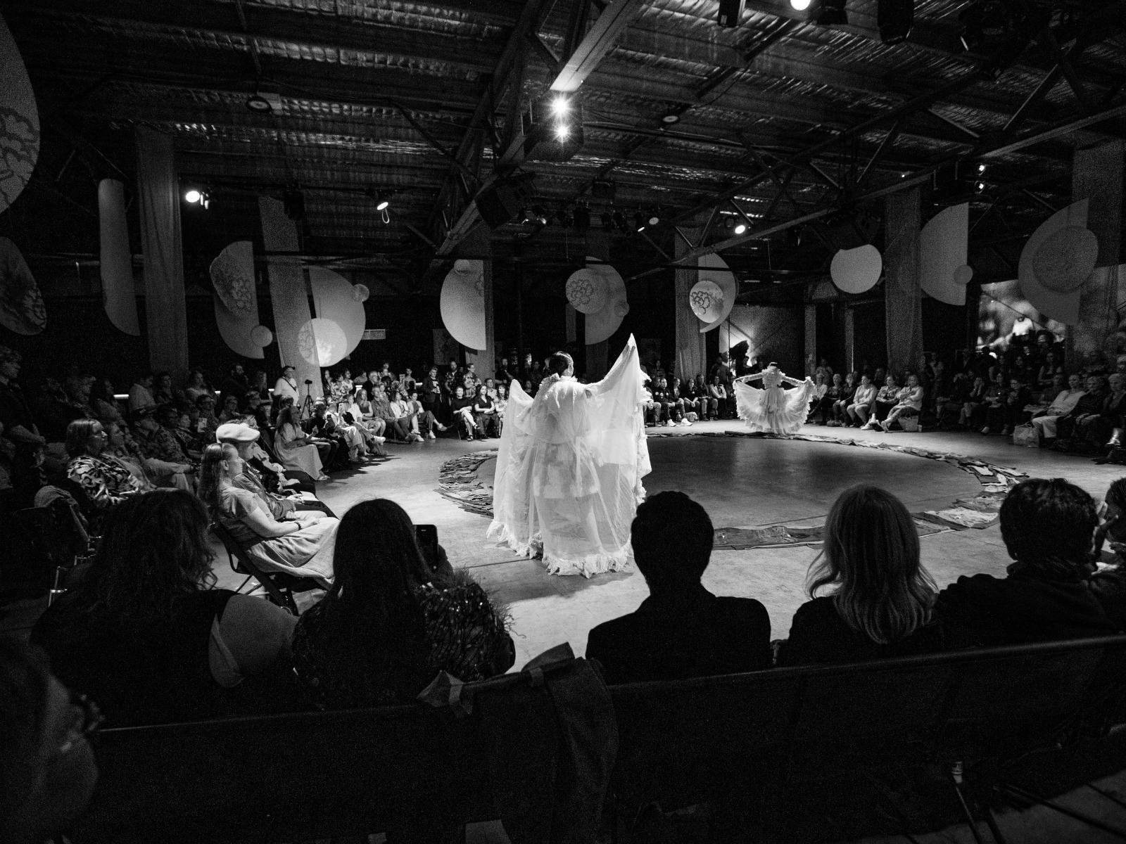 The audience sits in a large circle. In the middle, a figure in white holds a graceful pose.