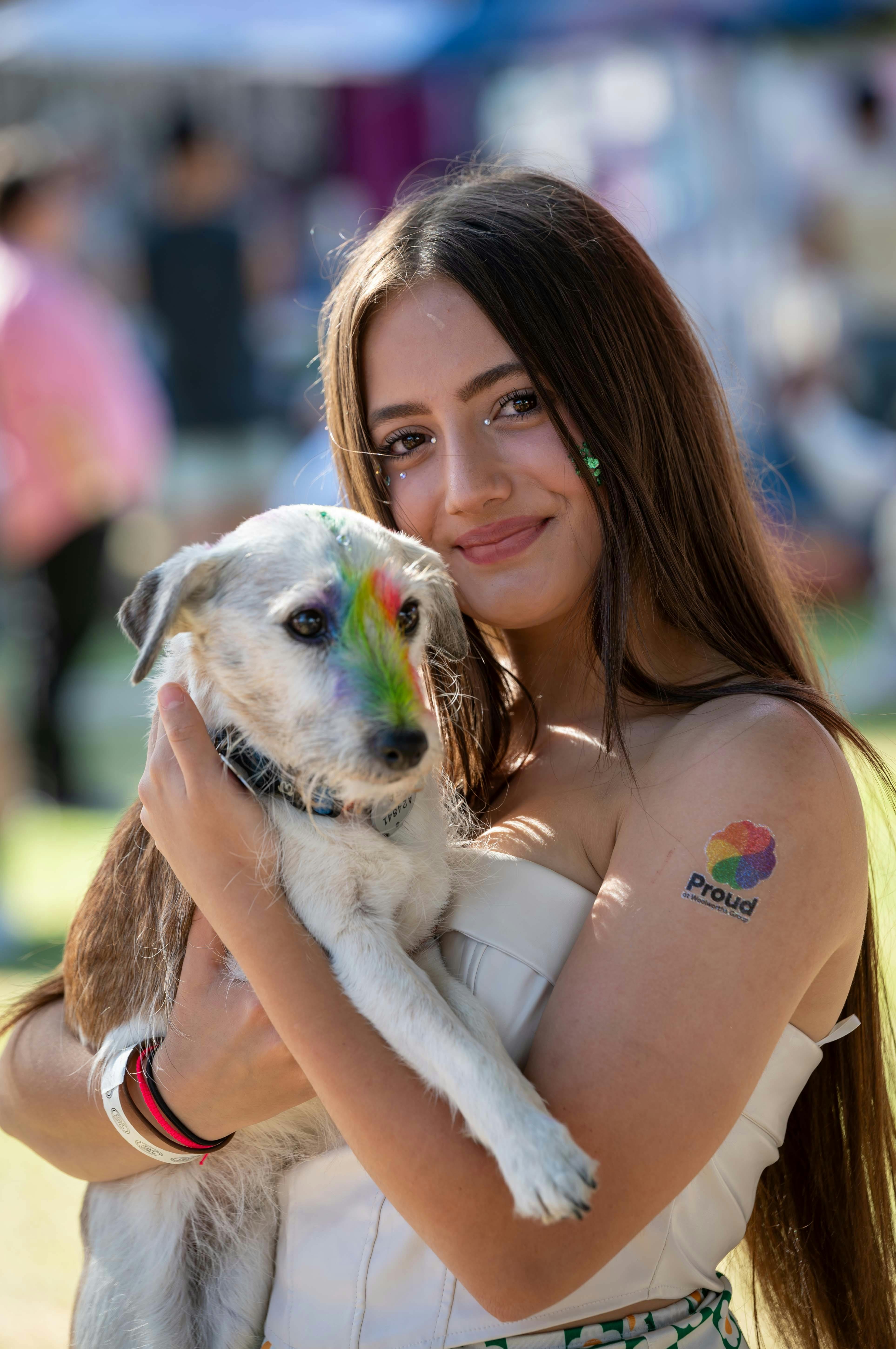 A person smiling and holding their small white dog with rainbow colours painted on their head.