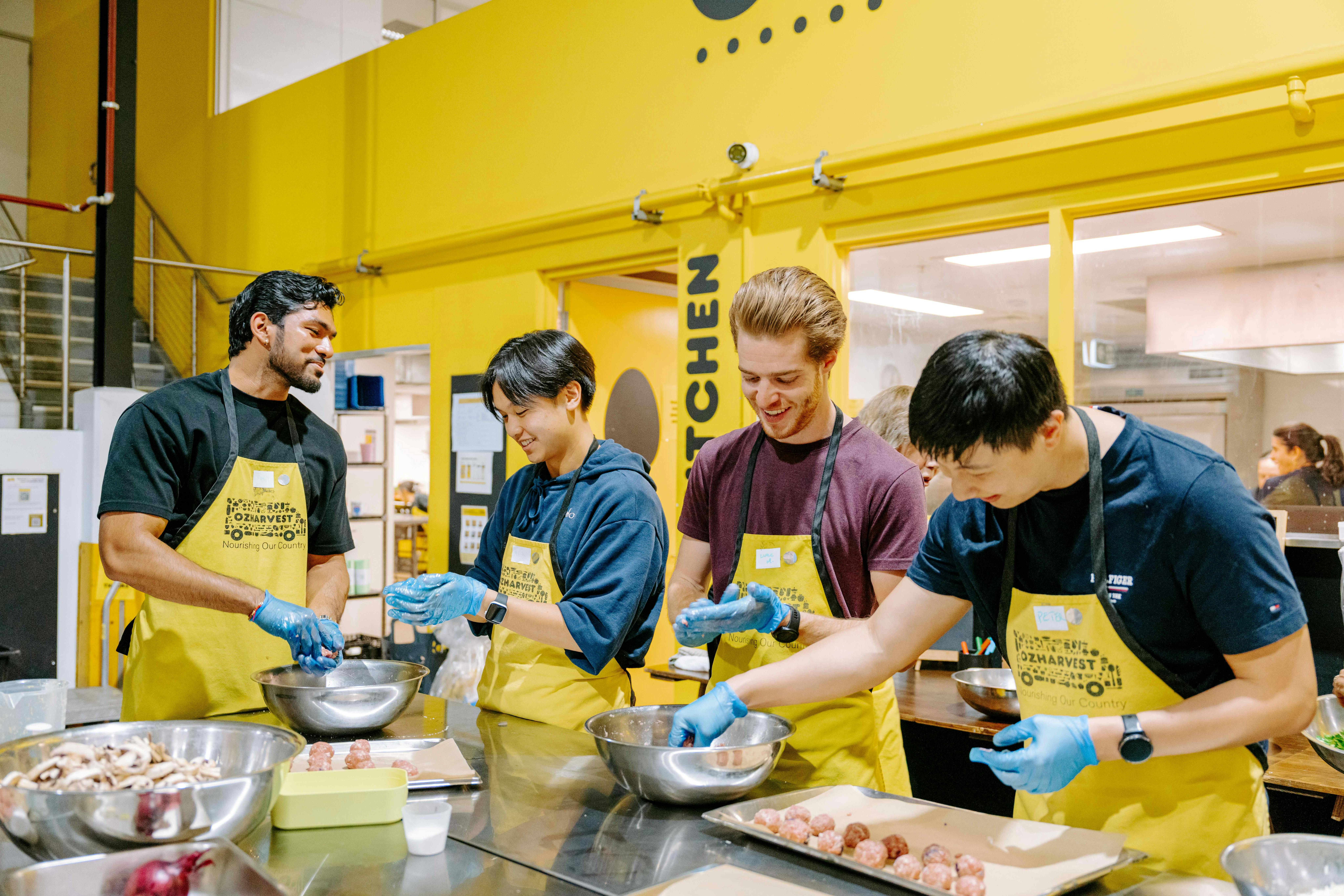 Four people rolling meatballs by hand during an OzHarvest cooking class, wearing gloves and yellow O
