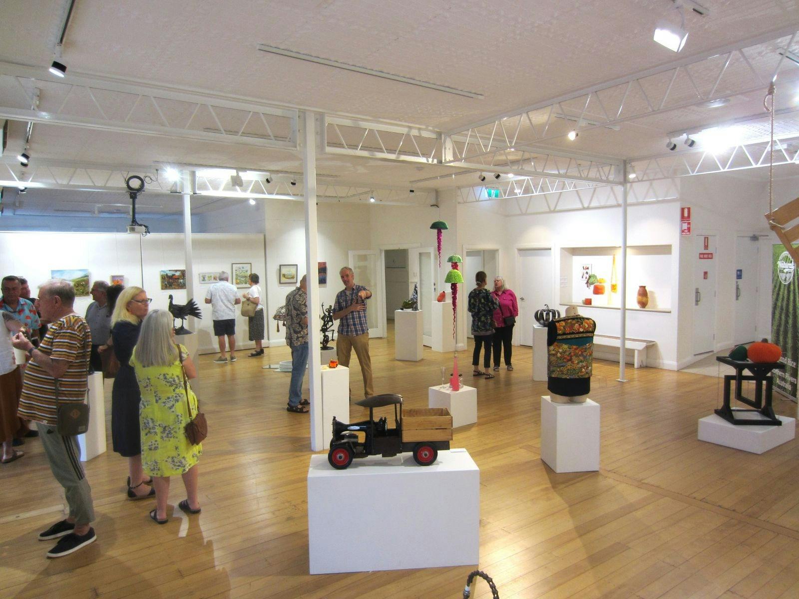 A room full of people talking and plinths with different types of craft on them.