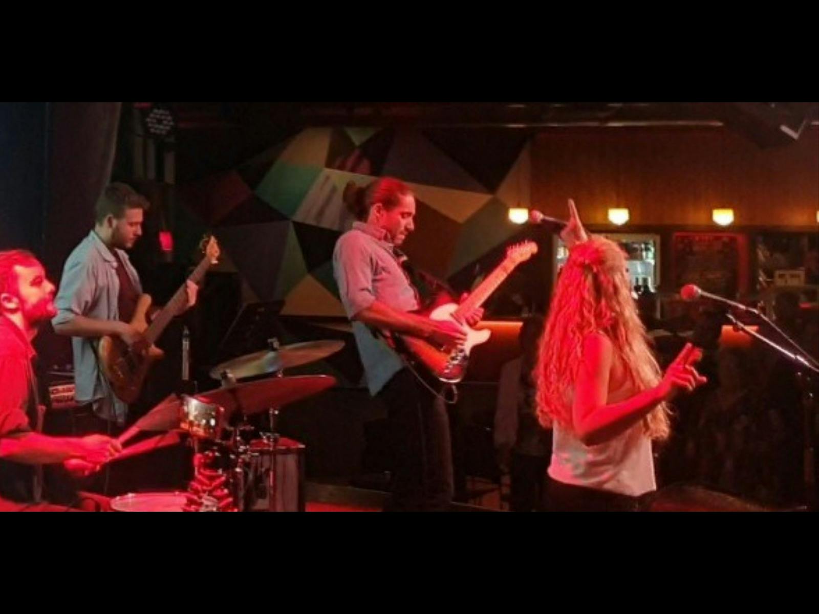 A live rock band performs in red stage lighting: a singer with long hair faces the crowd.